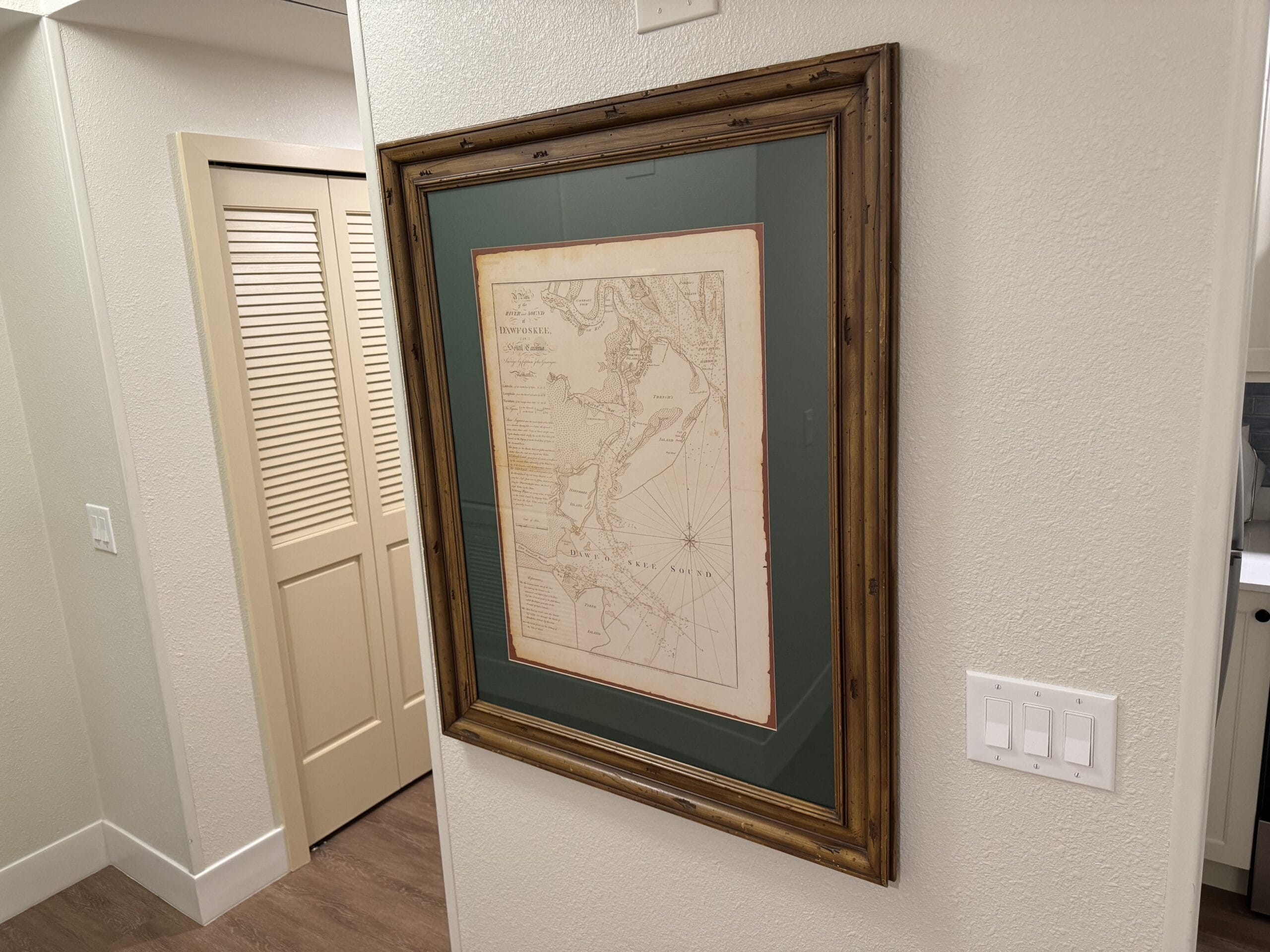 A framed historical map adorns the wall next to a light switch and a louvered closet door in a remodeled 1-bedroom villa at Disney's Hilton Head Island Resort, boasting light-colored walls and wooden flooring.