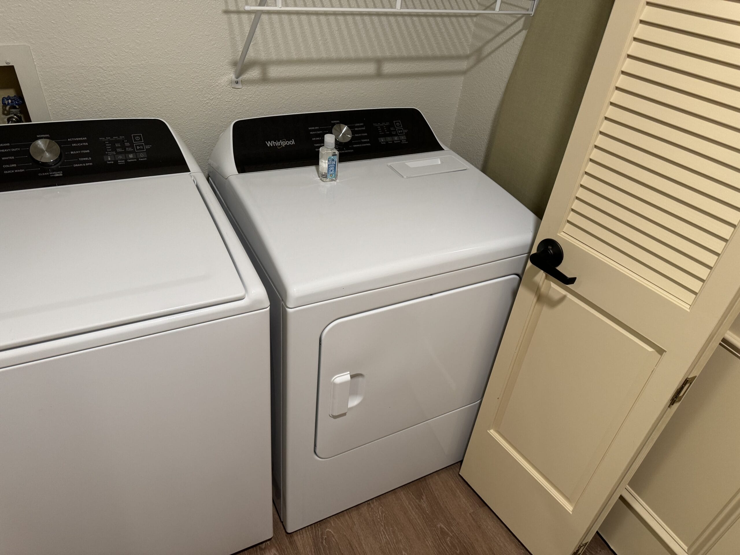 Step into the laundry room of this remodeled 1-bedroom villa at Disney's Hilton Head Island Resort. The washer on the left complements the dryer on the right, with a small bottle perched atop. Above, a wall-mounted shelf and louvered door complete this thoughtful design tour.