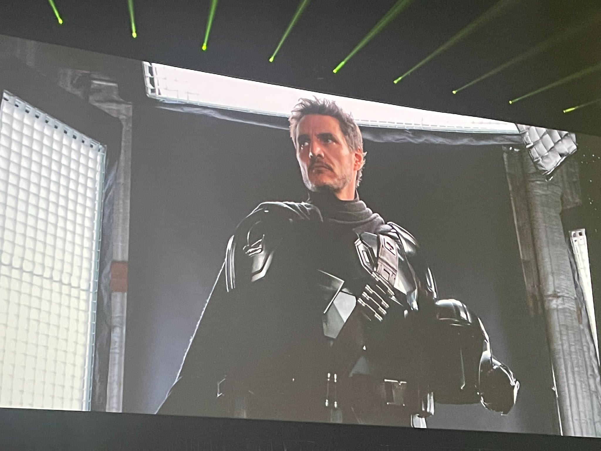 A man in futuristic black armor stands in a dimly lit room with industrial lighting panels on the walls and ceiling.