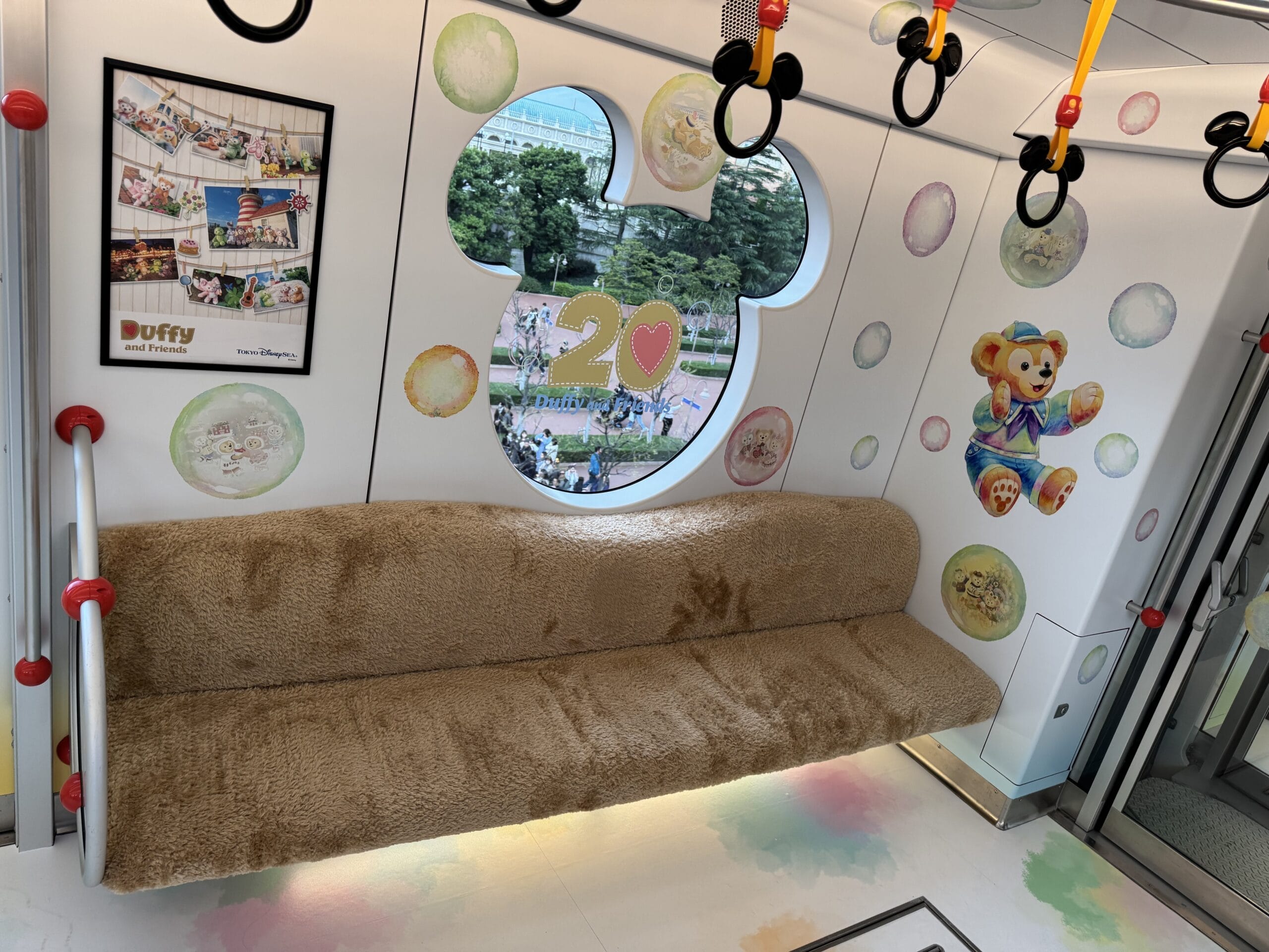 A cozy monorail interior at Tokyo Disney Resort features a fuzzy brown bench, Disney-themed decorations, a Mickey-shaped window with “20♥”, and charming Duffy & Friends cartoon bear illustrations on the walls.
