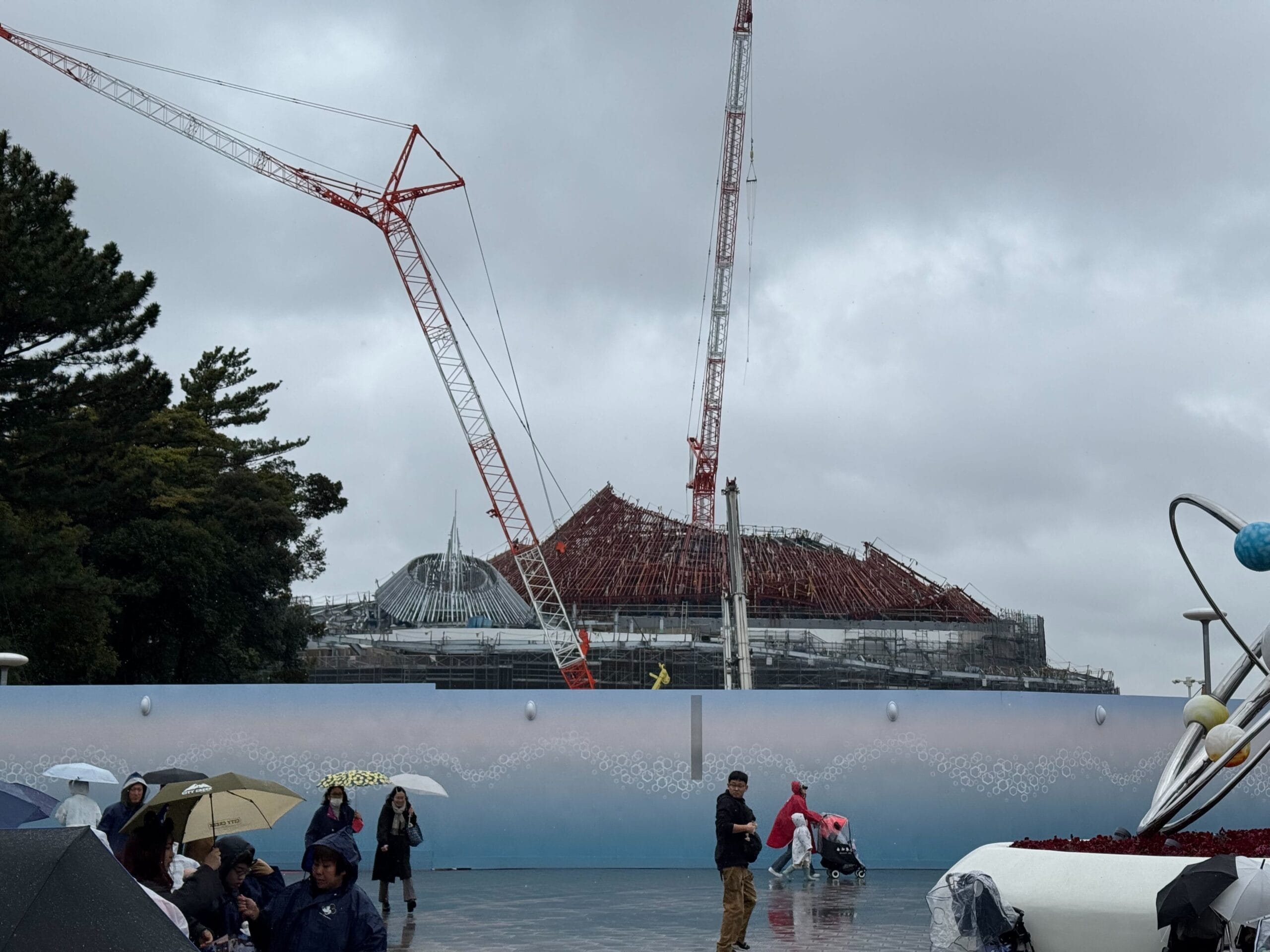 Construction site with a large dome structure being built using cranes. People with umbrellas walk by under overcast skies.