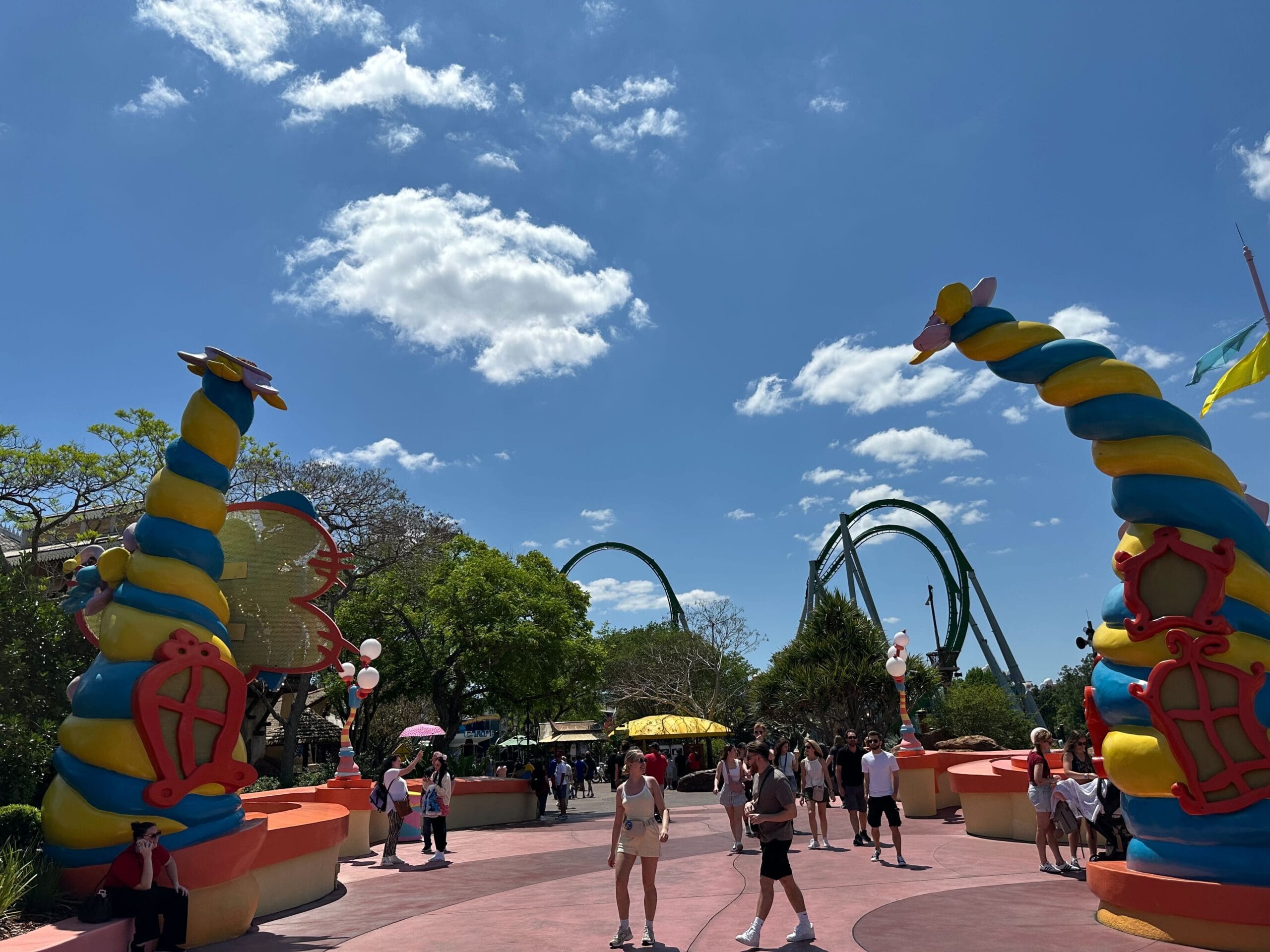 Amusement park scene with colorful dragon sculptures and roller coaster tracks in the background. Visitors walk along a sunny path under a blue sky with scattered clouds.