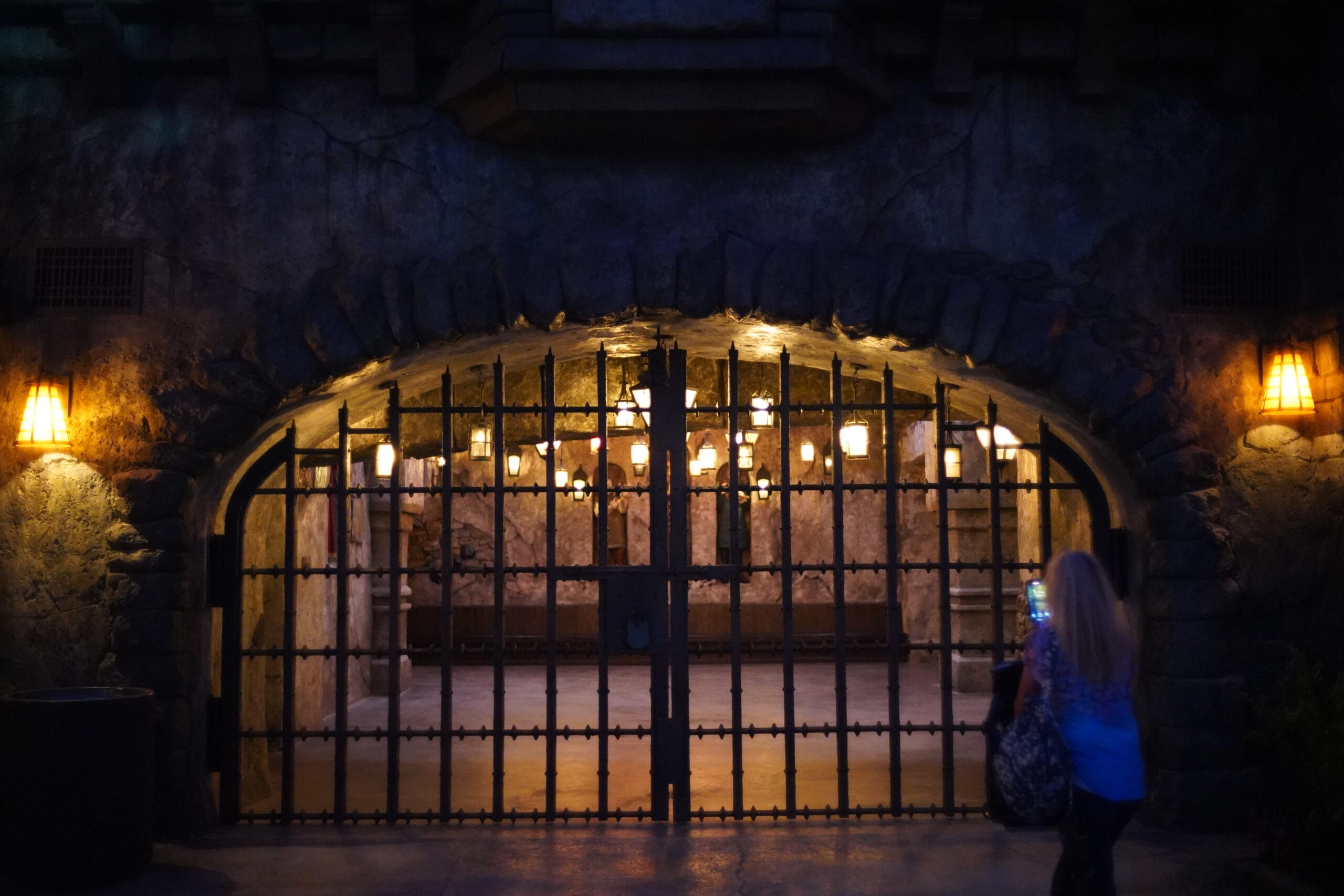 A woman stands near an iron gate with warm lights in Das Stakehaus, surrounded by stone walls in a castle-like setting.