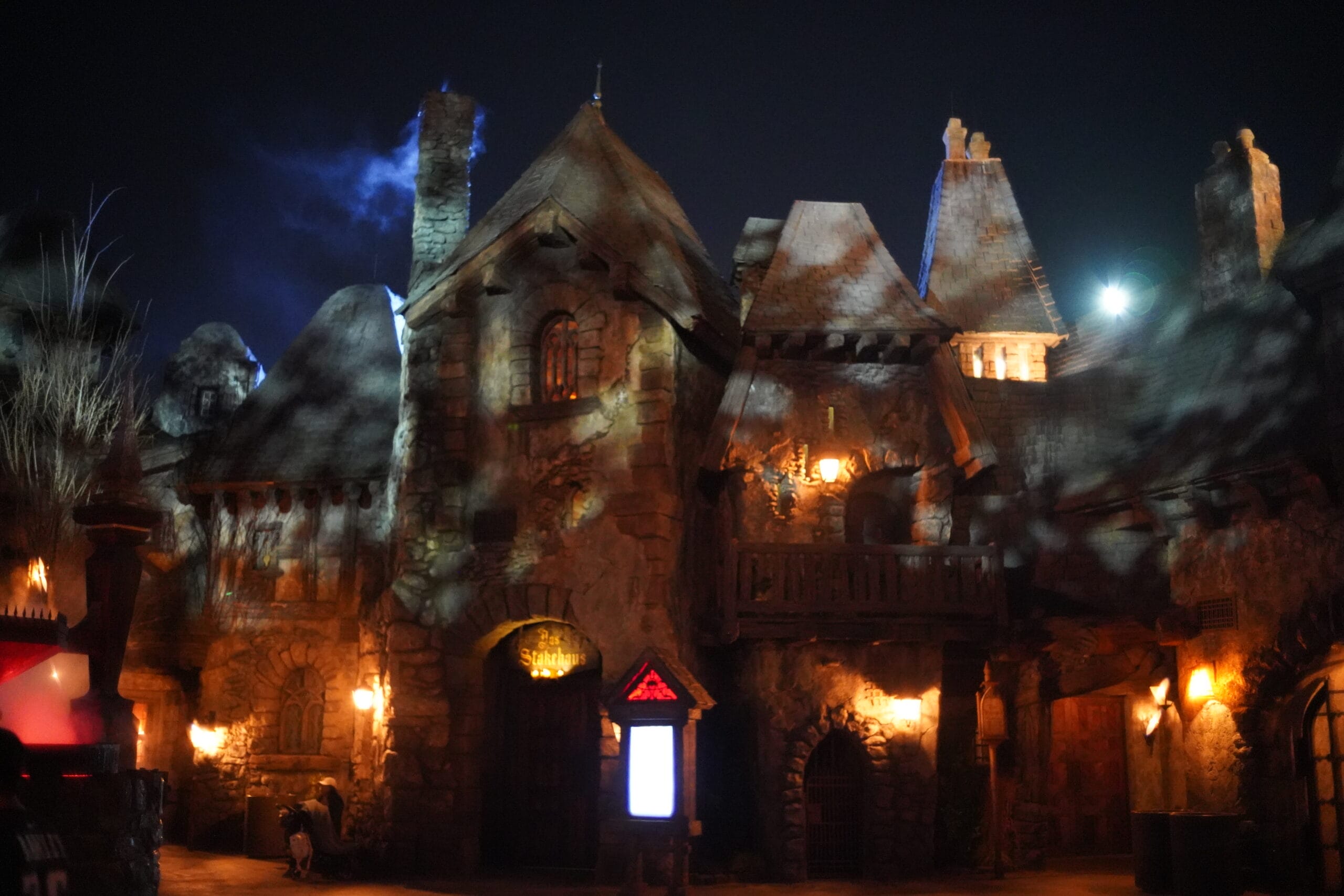 A spooky, dimly lit stone castle—Das Stakehaus—with pointed roofs, aglow in eerie orange and blue lights at night.