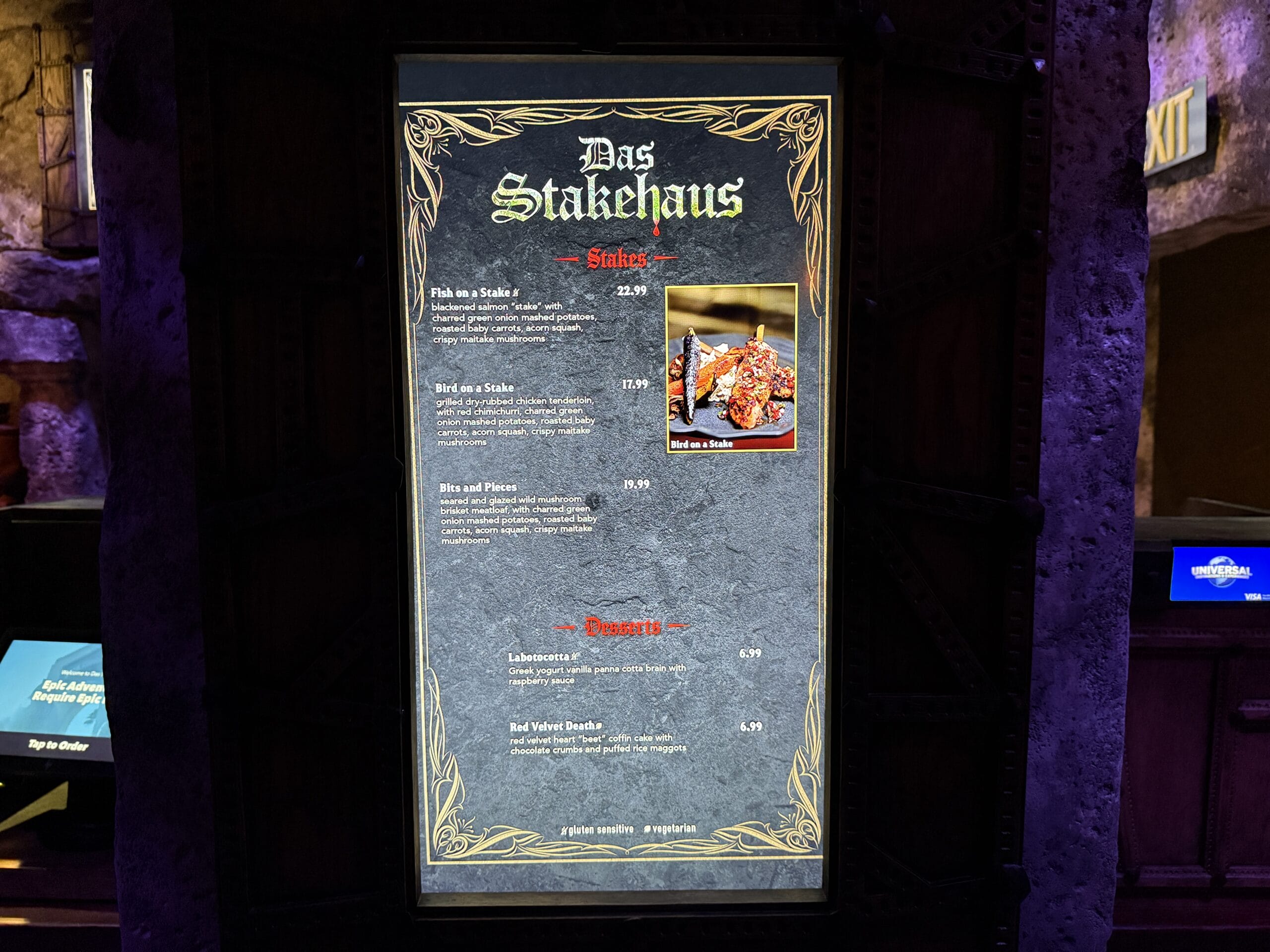 Das Stakehaus illuminated menu board with gothic design displays steaks, bites, pizzas, and desserts in a dark setting.
