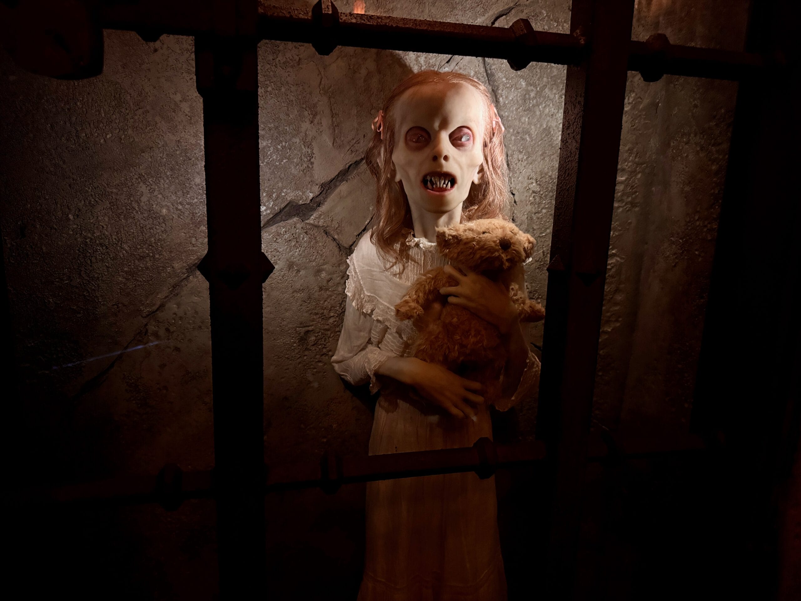 A creepy Das Stakehaus doll with sharp teeth and wide eyes grips a teddy bear behind metal bars in a dimly lit room.