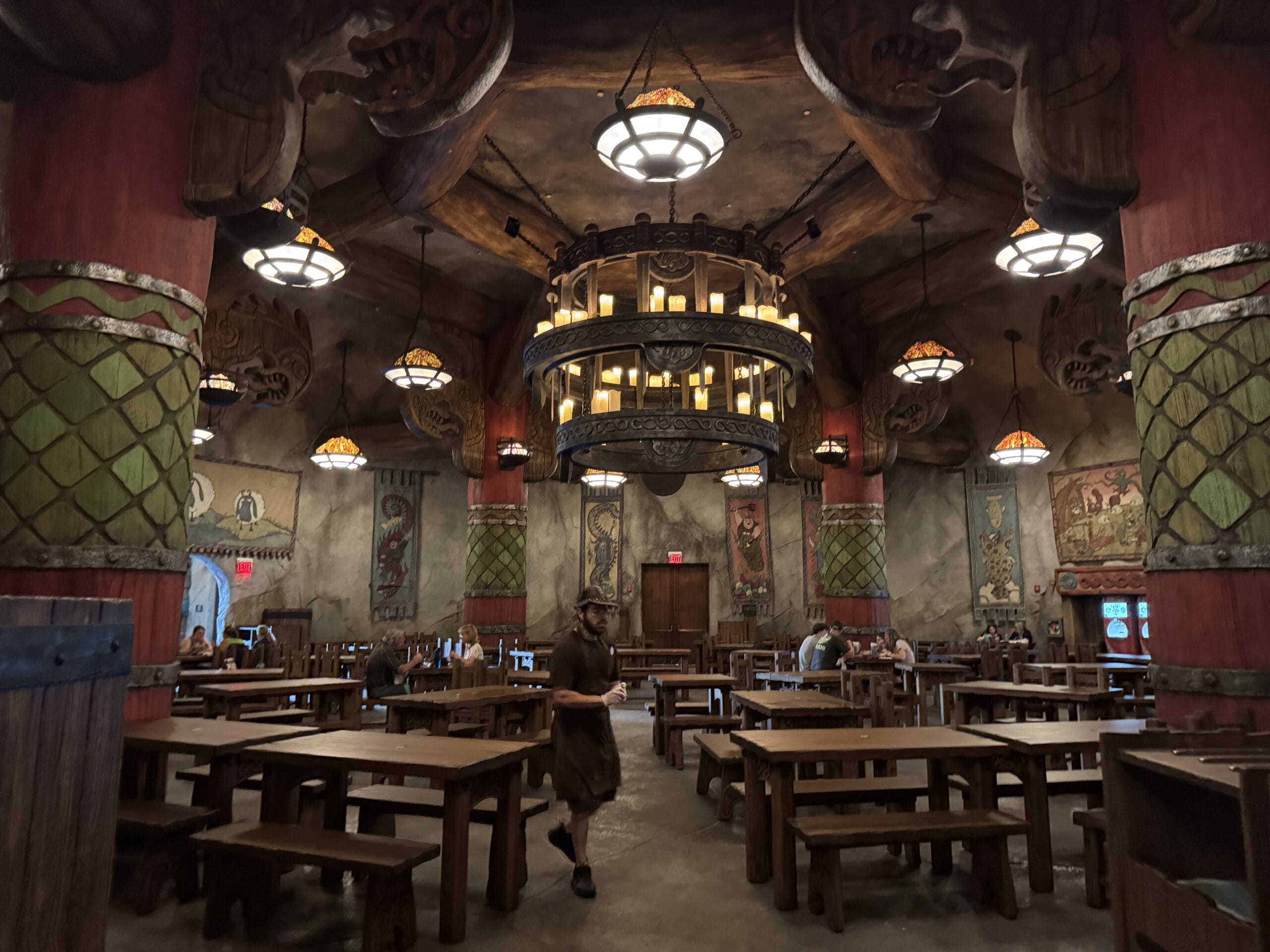 A large Mead Hall with wooden tables and benches, Viking-inspired decor, hanging lanterns, and a few people seated and walking inside.