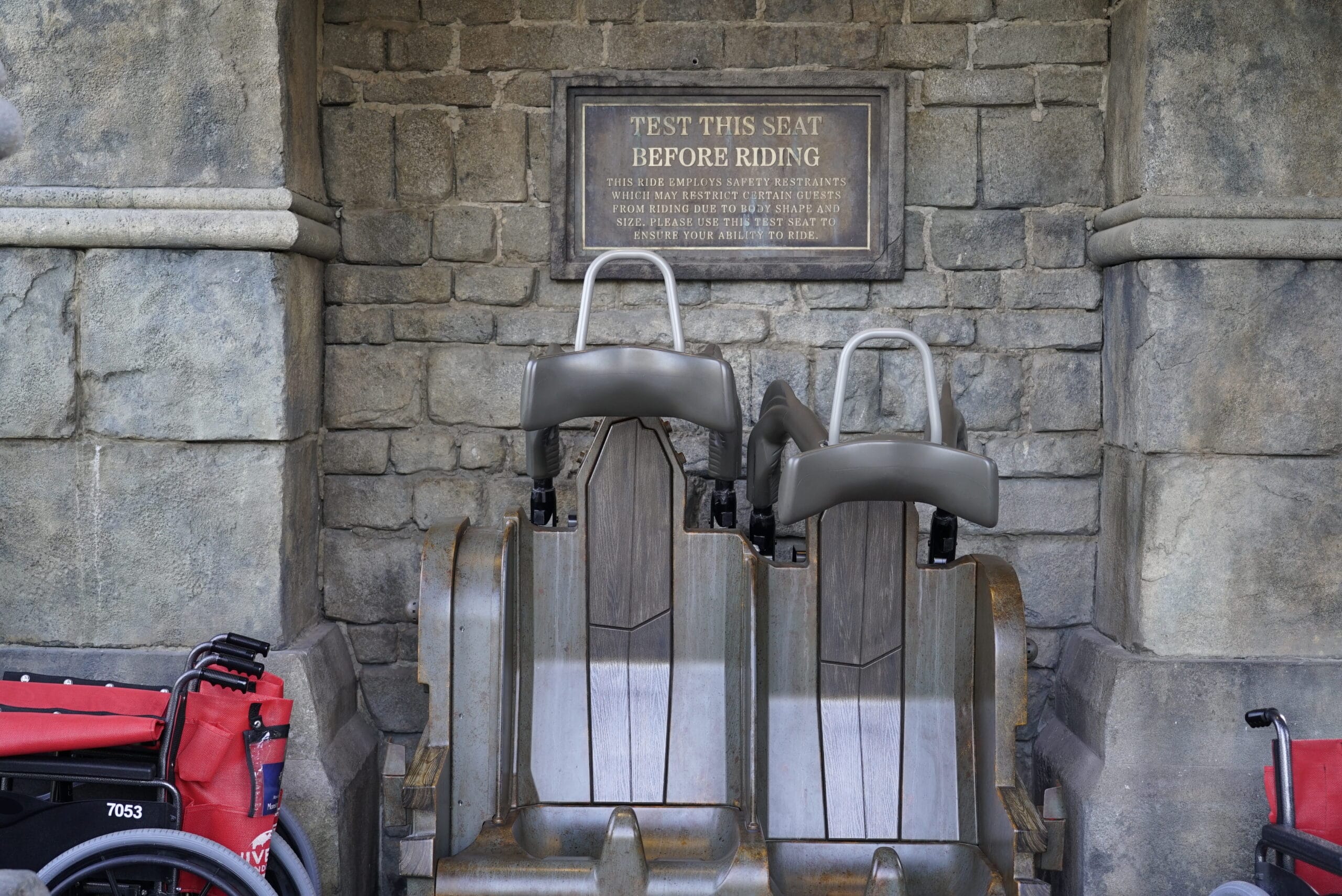 Two amusement park ride seats with safety restraints sit under a sign reading "Test This Seat Before Riding," against a stone wall—just steps from the Monsters Unchained: The Frankenstein Experiment attraction and two red mobility scooters.