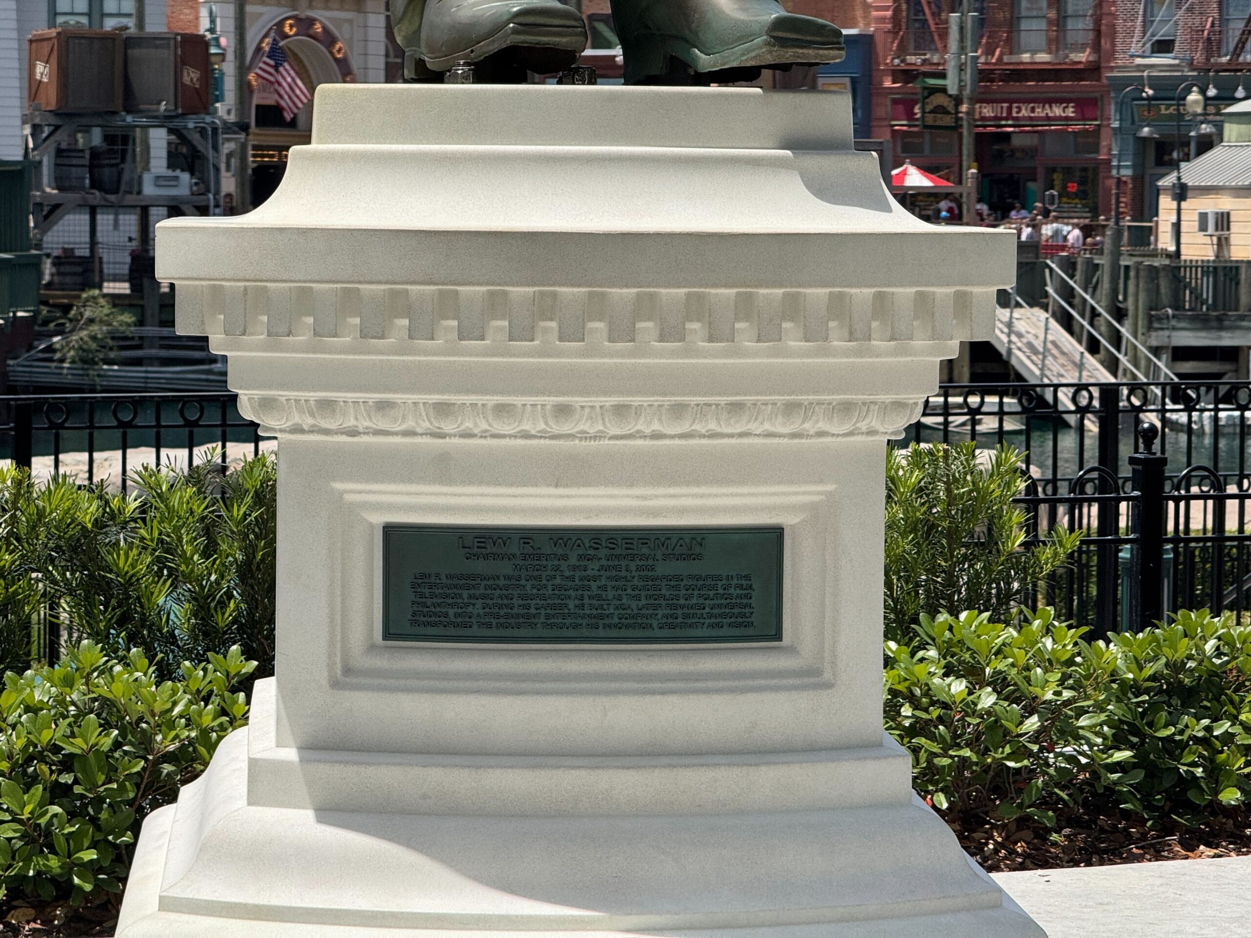 White stone pedestal with engraved plaque, topped with a statue of two feet wearing shoes, outdoors near buildings.