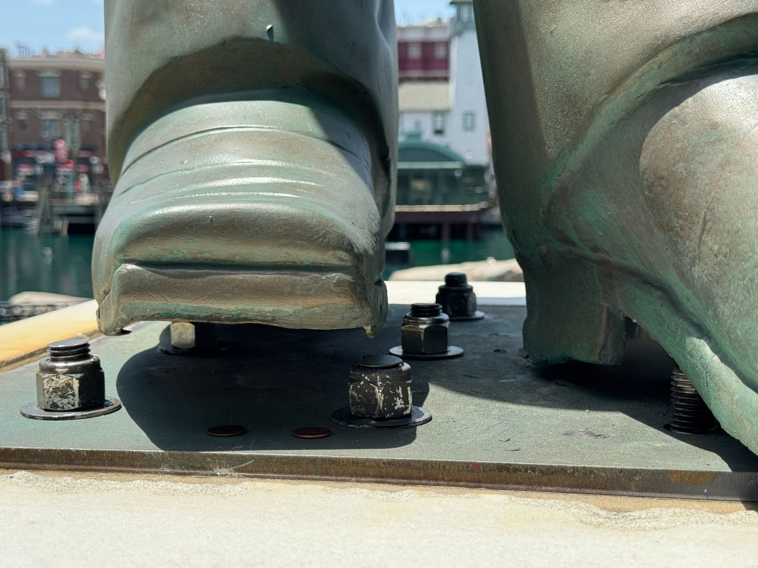 Close-up of a statue’s feet on a metal base secured with bolts, with two pennies lying near the left boot.