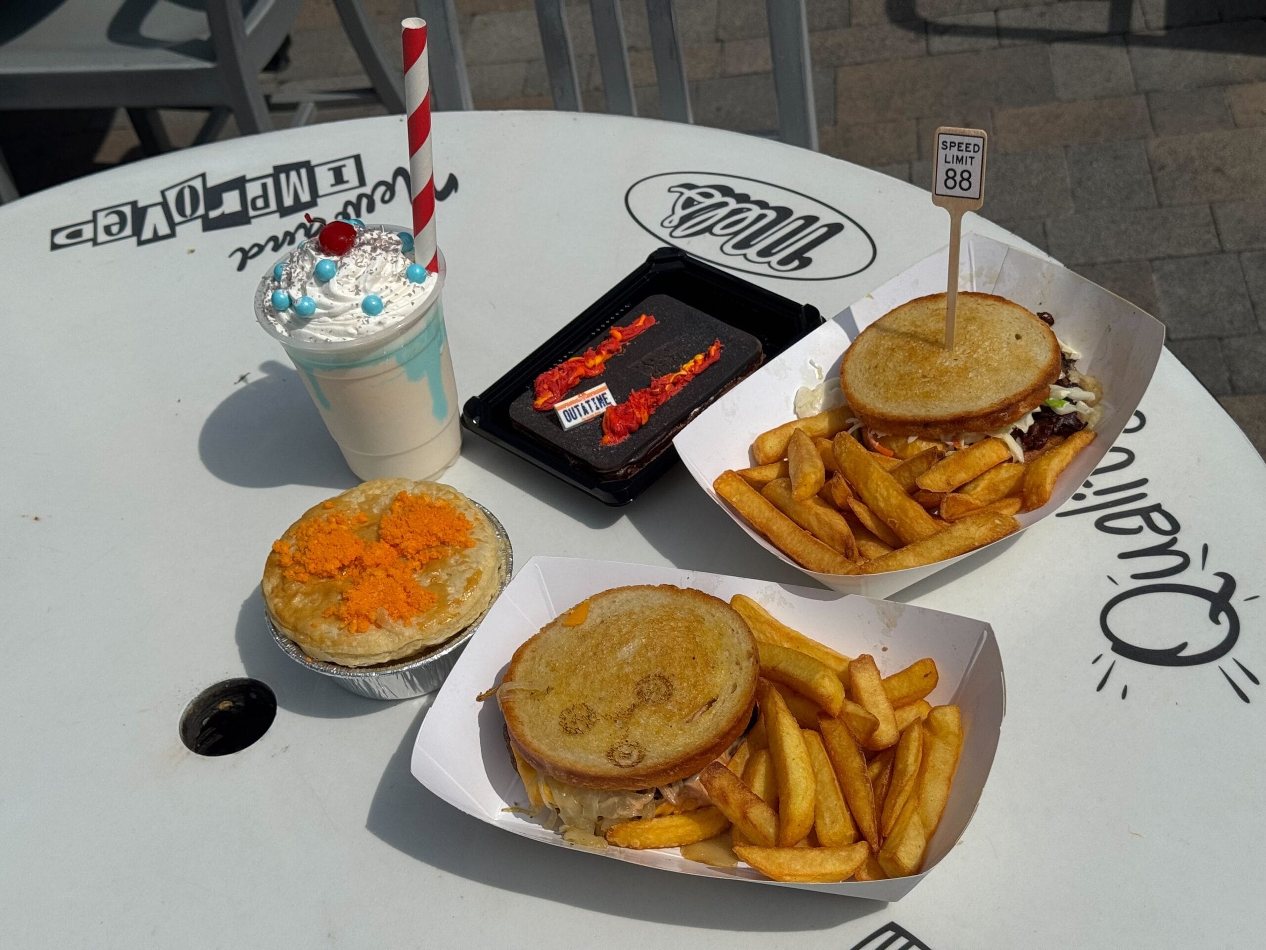 Two trays with burgers and fries, a pie, a milkshake with whipped cream, and a brownie sit on a white outdoor table.