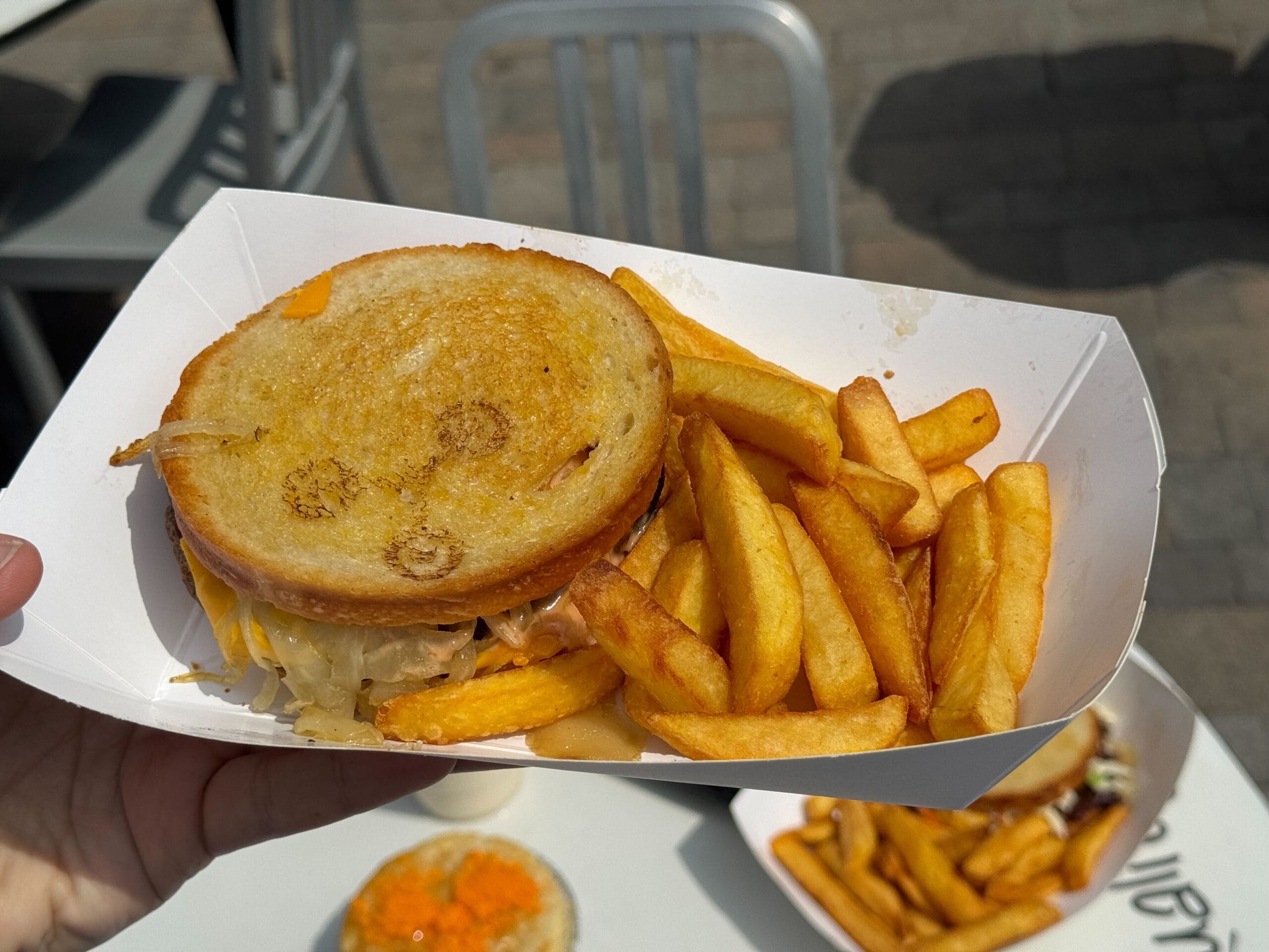 A hand holds a tray with a grilled sandwich and golden fries; outdoor seating is visible in the background.