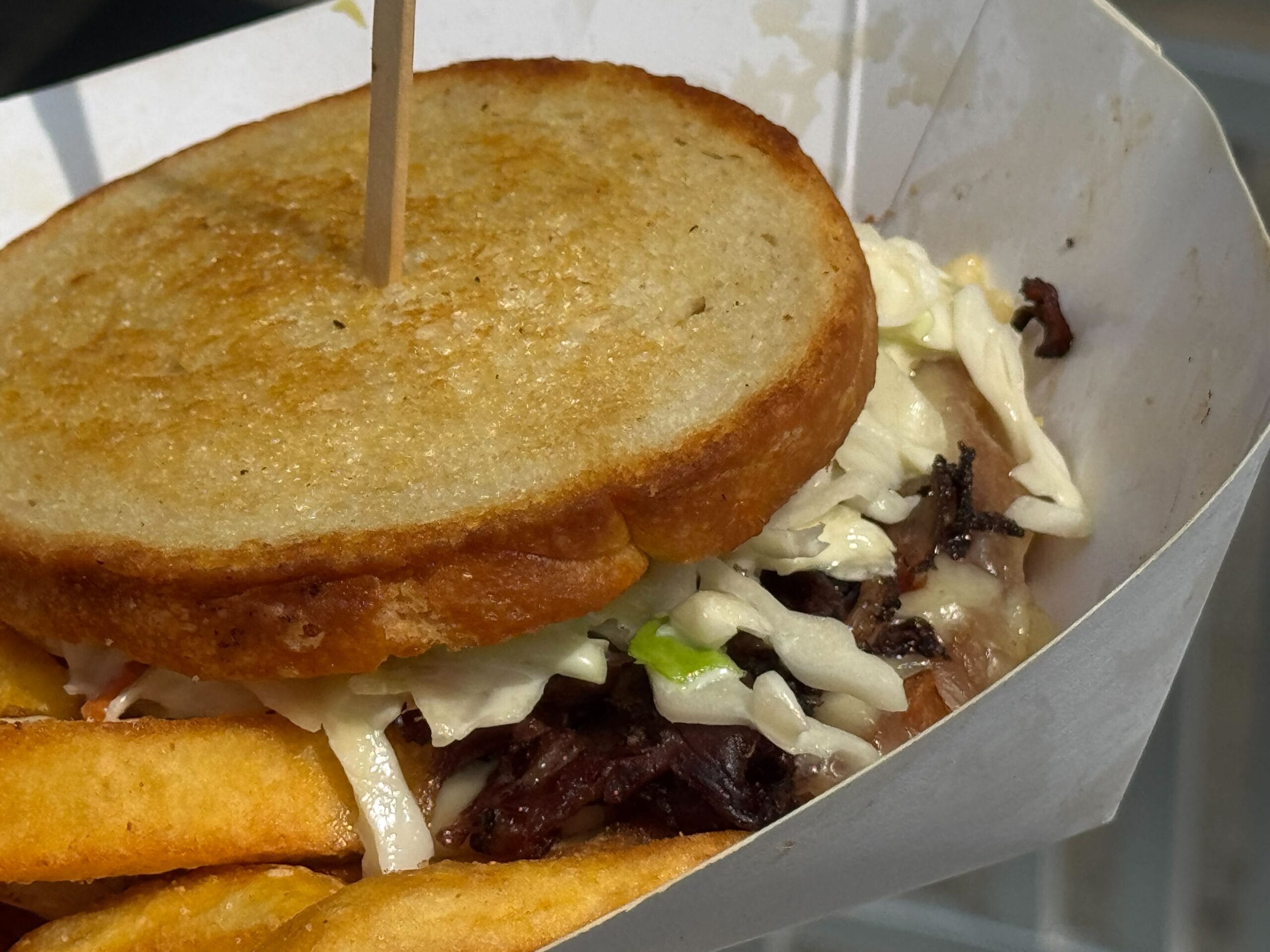A close-up of a bttf food sandwich with coleslaw and fries in a paper tray, secured with a wooden pick.