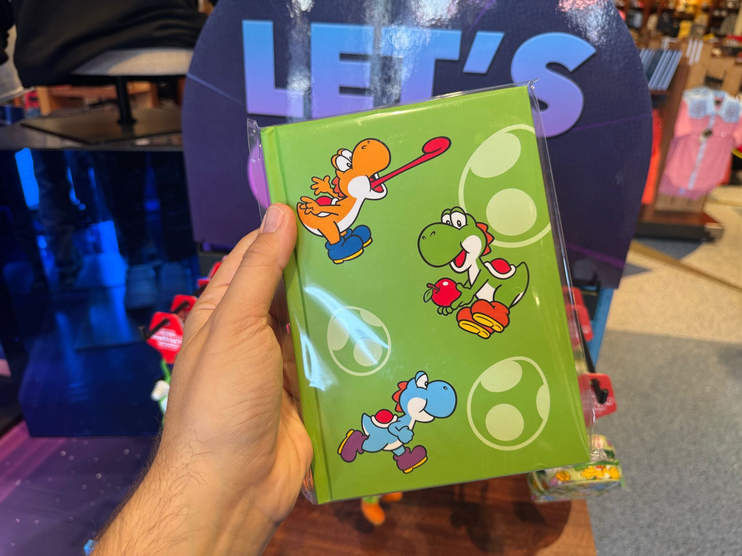 A hand holds Yoshi merchandise: a green notebook with colorful Yoshi characters and eggs from the Mario series on the cover.