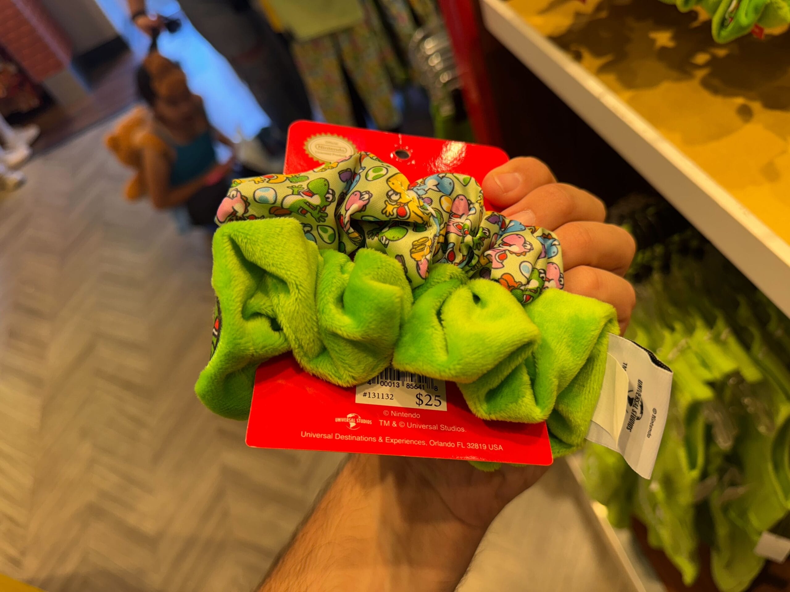 A hand holds Yoshi merchandise—green, cartoon-patterned scrunchies—on a red card in a Universal Studios Hollywood store.