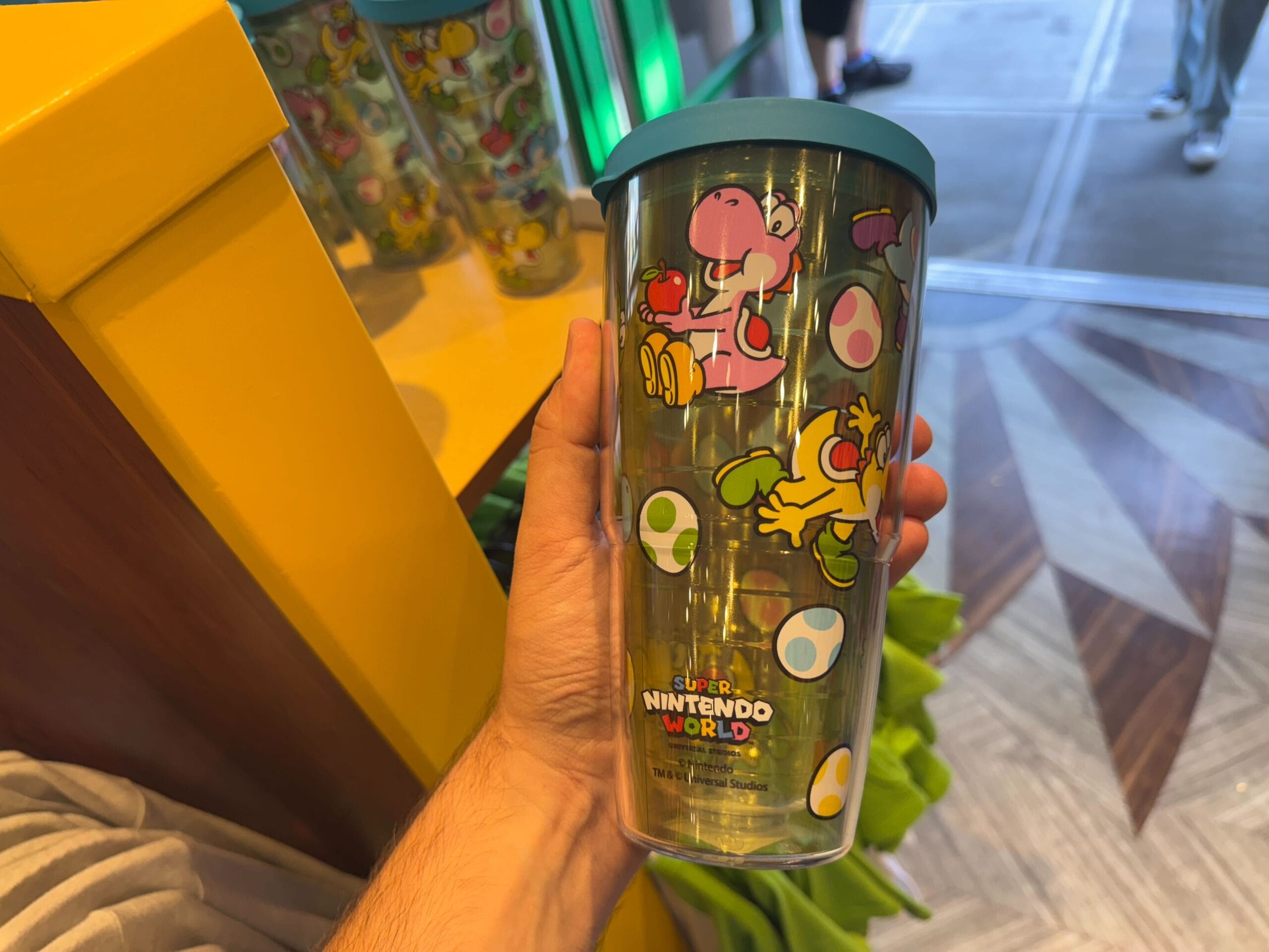 Hand holding Yoshi merchandise tumbler with bright designs at Universal Studios Hollywood display shelf.