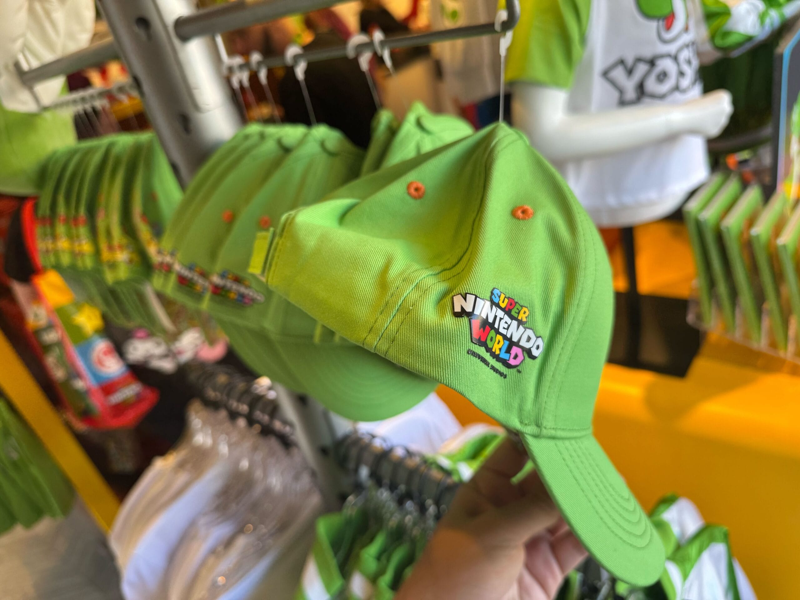 A green Super Nintendo World cap is held in a store, surrounded by Yoshi merchandise at Universal Studios Hollywood.