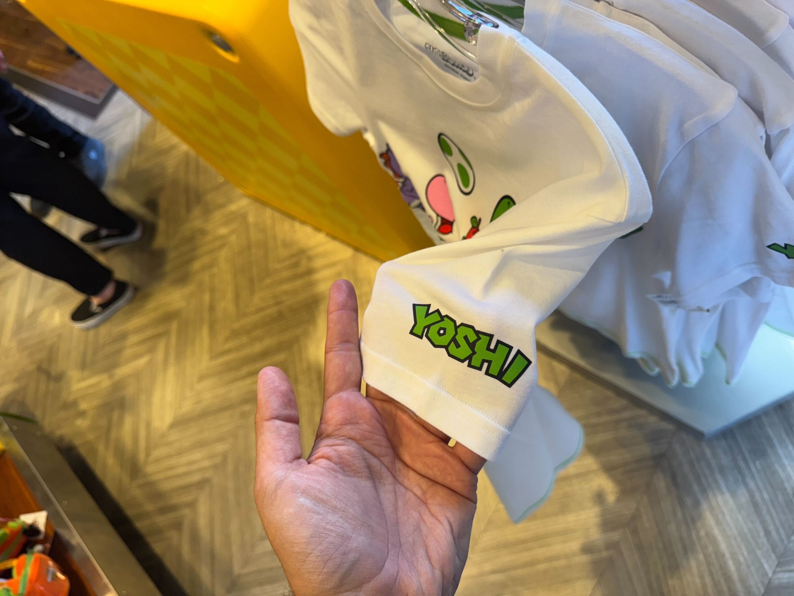 A hand holds a white Yoshi merchandise sleeve at Universal Studios Hollywood, with shirts on display for Fan Fest Nights.