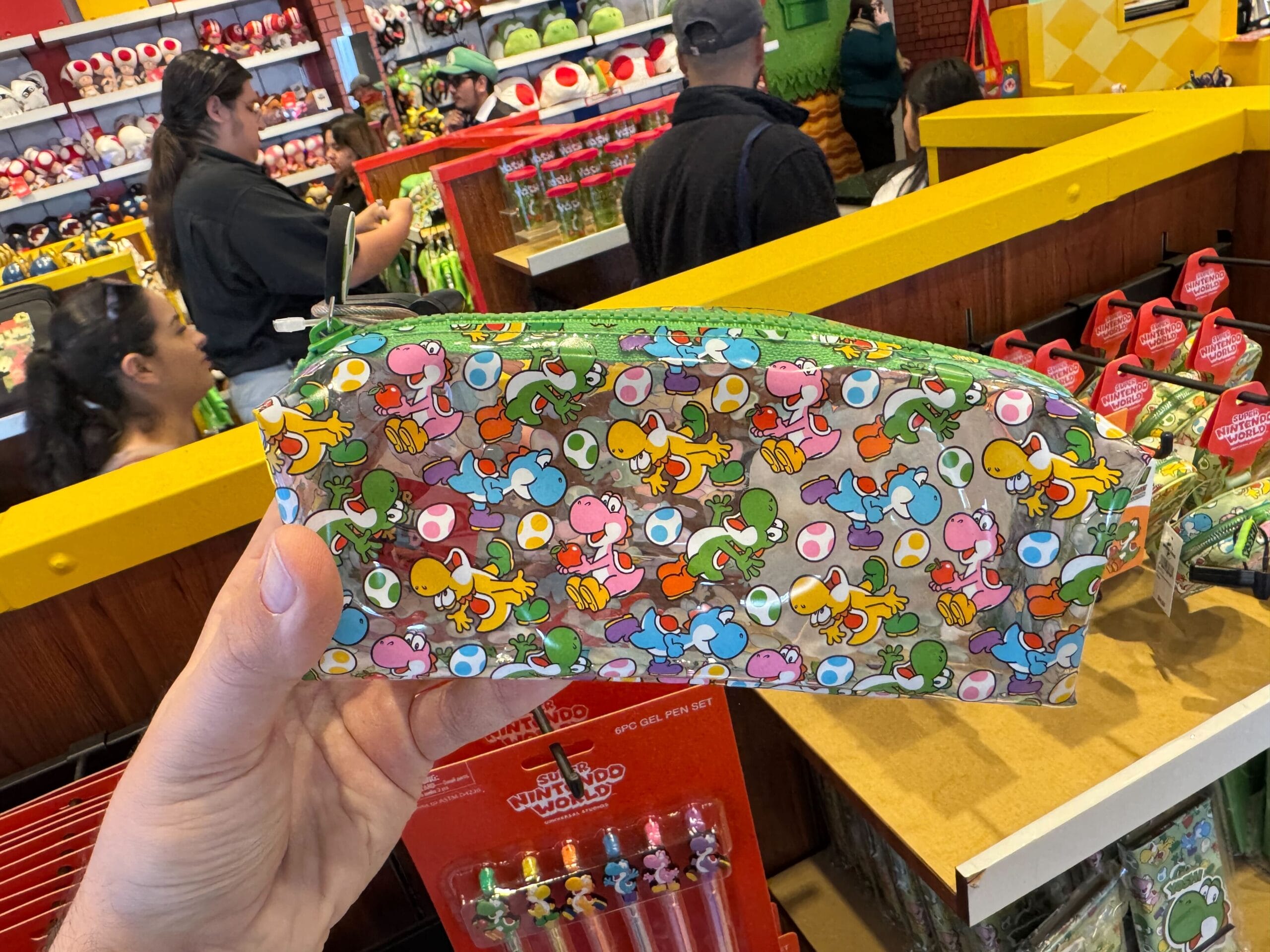 A hand holds Yoshi merchandise—a colorful pencil case—in a busy Universal Studios Hollywood store filled with shoppers.