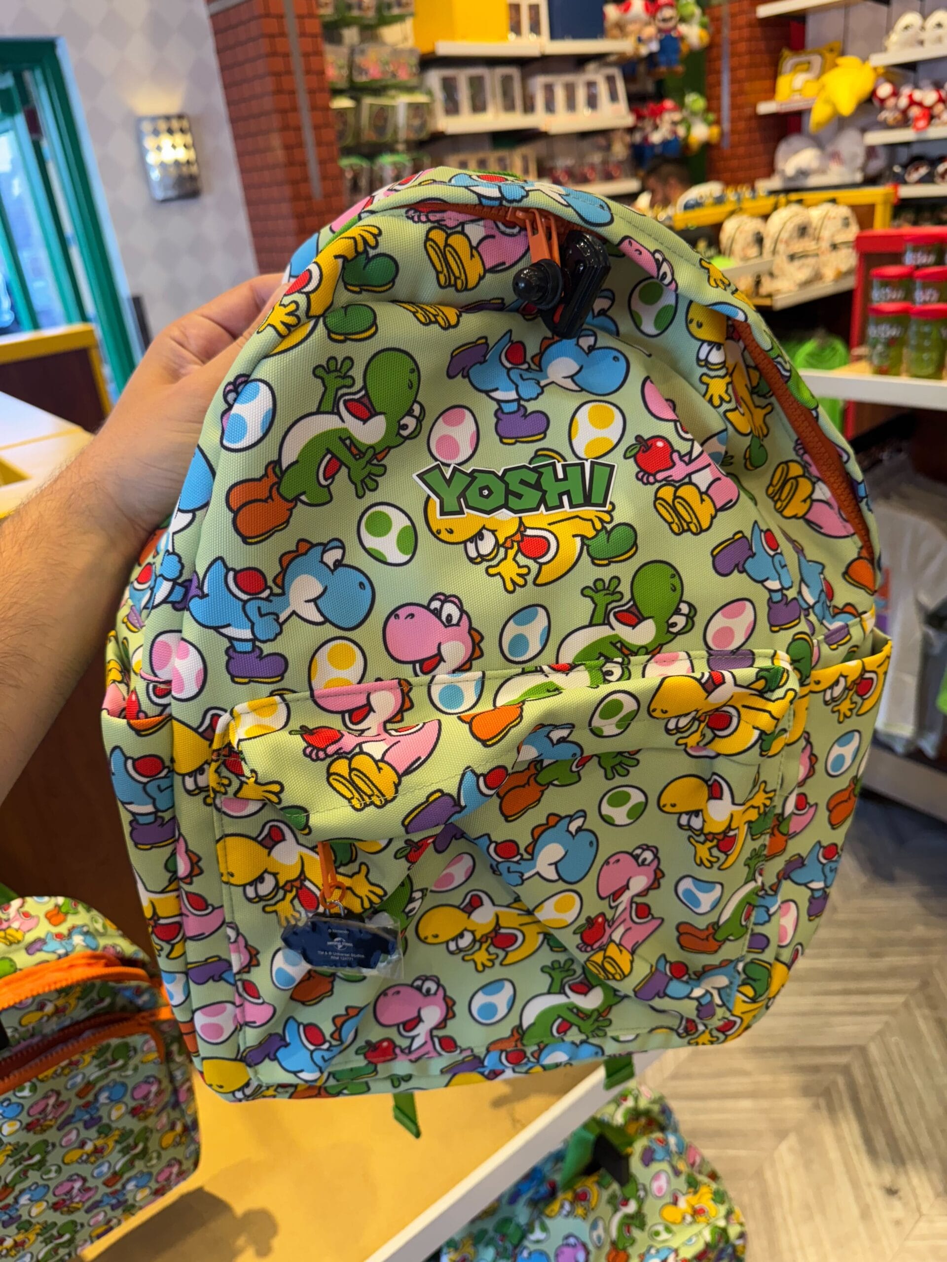 A hand holds a colorful Yoshi-themed backpack, perfect Yoshi merchandise for Universal Studios Hollywood fans.
