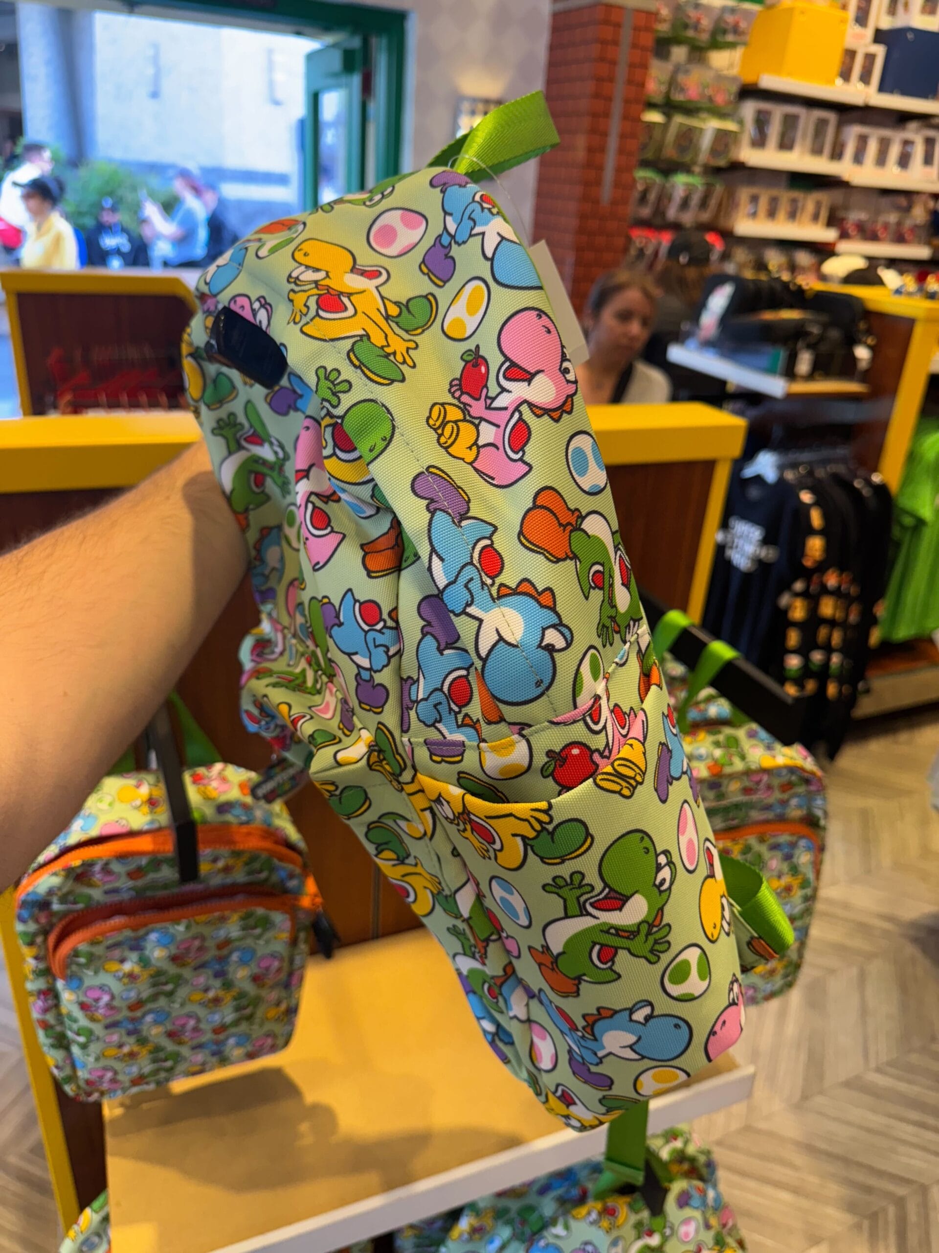 A hand holds a colorful Yoshi merchandise backpack in a Universal Studios Hollywood store with similar bags on display.