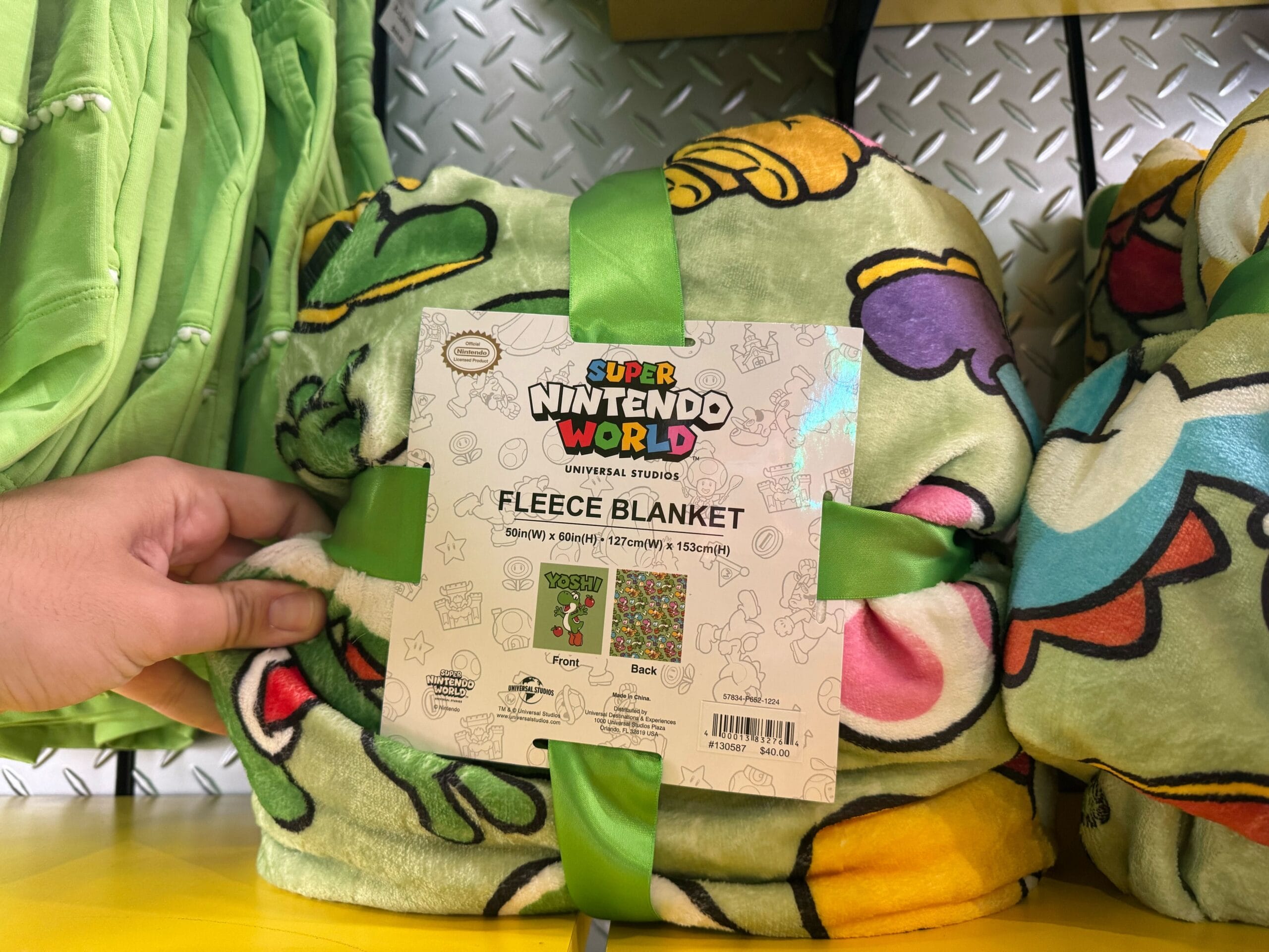 A hand holds Yoshi merchandise—a Super Nintendo World fleece blanket with cartoon characters and a green ribbon on display.