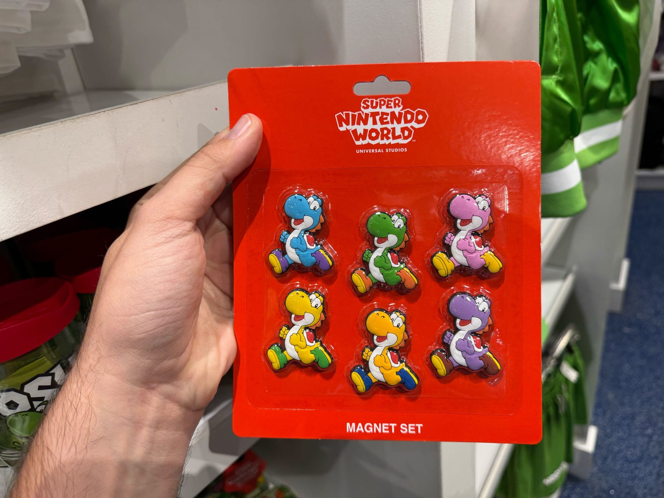 A hand holds Yoshi merchandise—six colorful Yoshi magnets from Universal Studios Hollywood—on a red backing card.