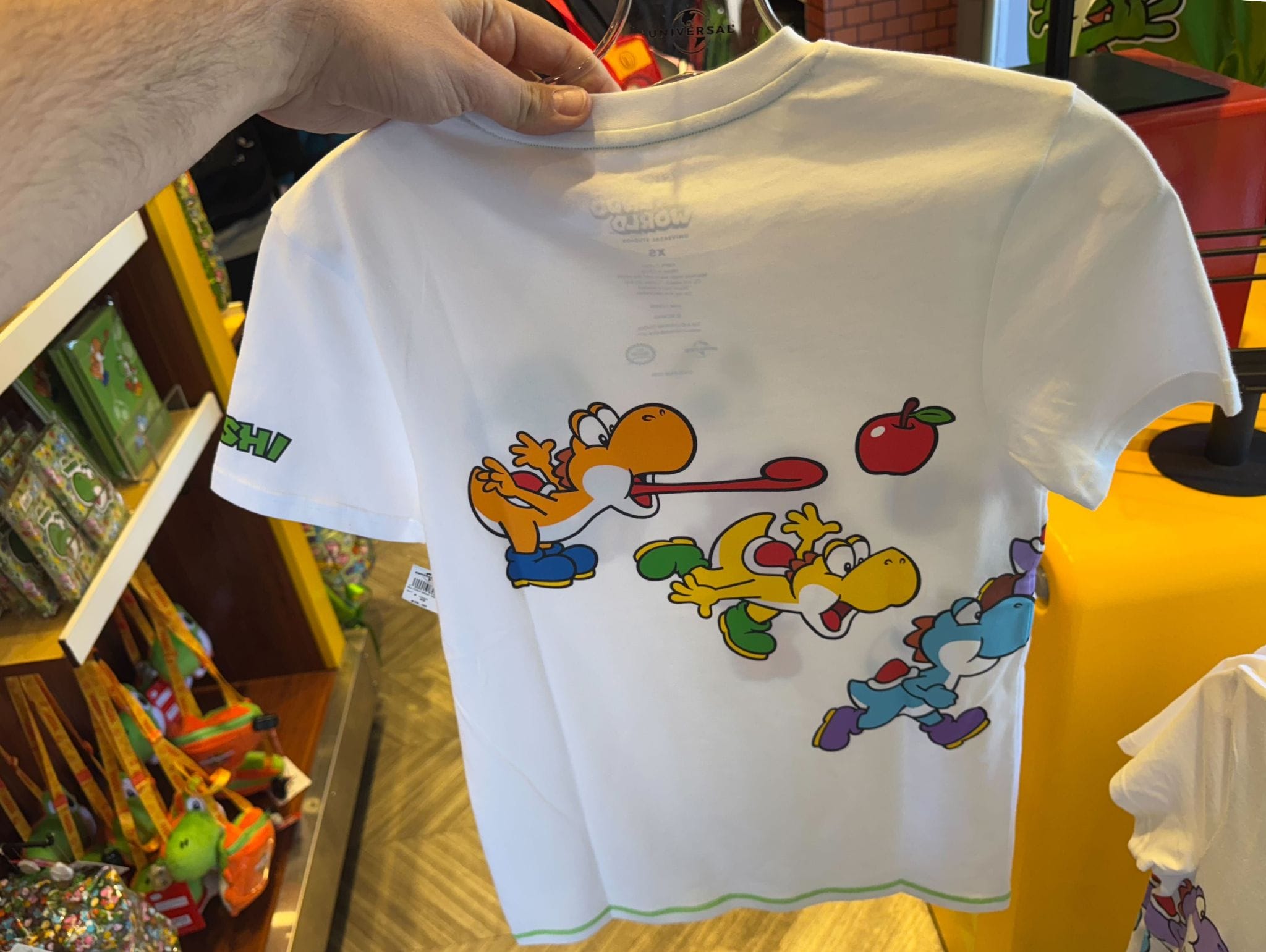 A hand holds Yoshi merchandise—a white tee with colorful Yoshis chasing an apple—perfect for Fan Fest Nights at Universal Studios Hollywood.