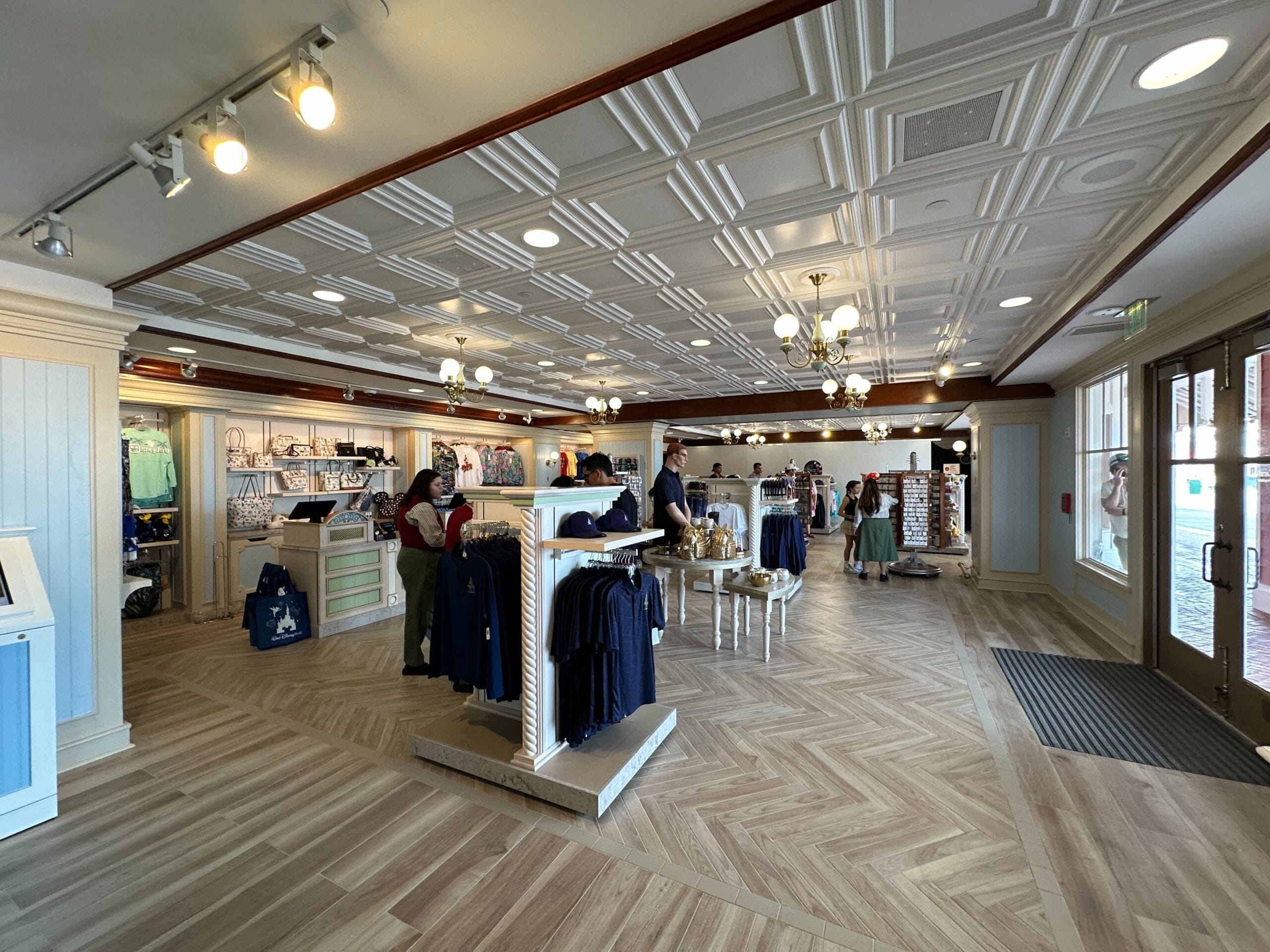 A retail store interior resembling Disney's Boardwalk Inn Resort features wooden floors and a patterned ceiling. Clothing displays and fixtures are arranged throughout, with people browsing the merchandise. Bright lights illuminate this beautifully renovated section, evoking charm and elegance.