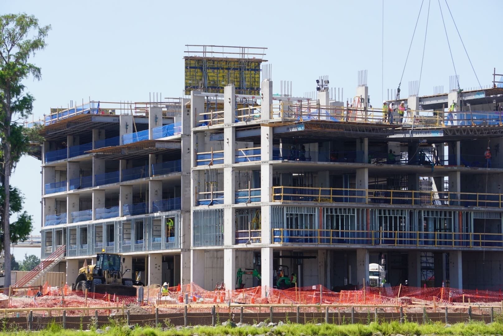 A multi-story building rises near the lakeshore lodge, adorned with scaffolding and safety netting. Construction equipment hums while workers diligently advance the project, blending modern architecture with nature's serene backdrop.