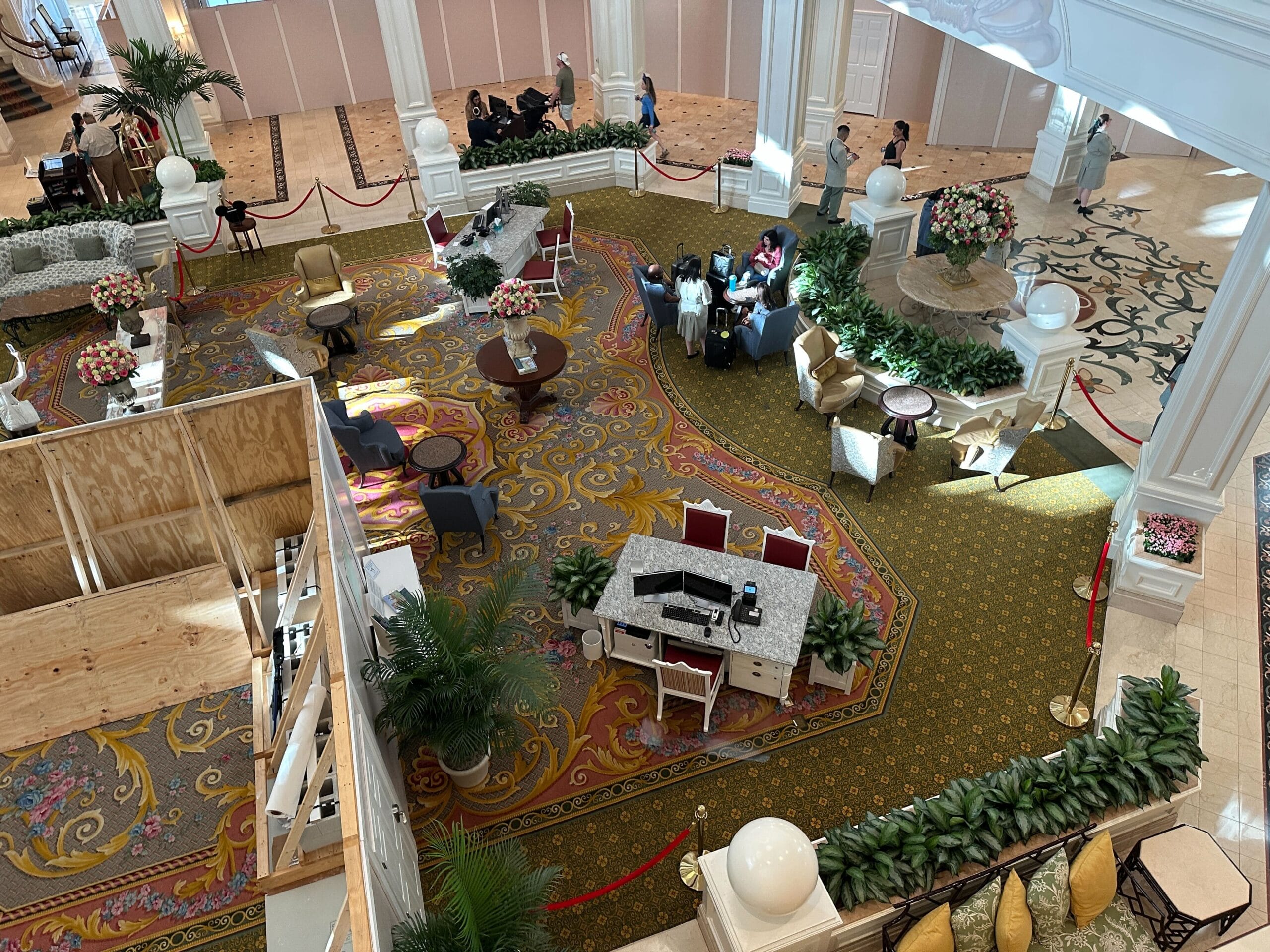 Hotel lobby with ornate carpet, flower arrangements, seating areas, and a construction barrier, viewed from above.