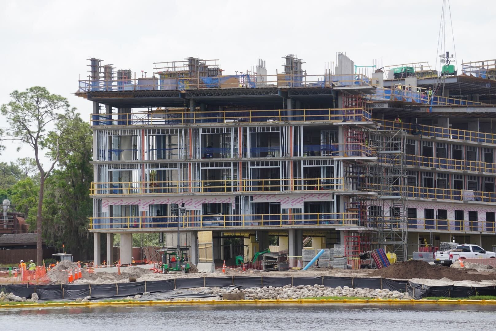 A multi-story building under construction beside water, with scaffolding, workers, and construction materials visible.