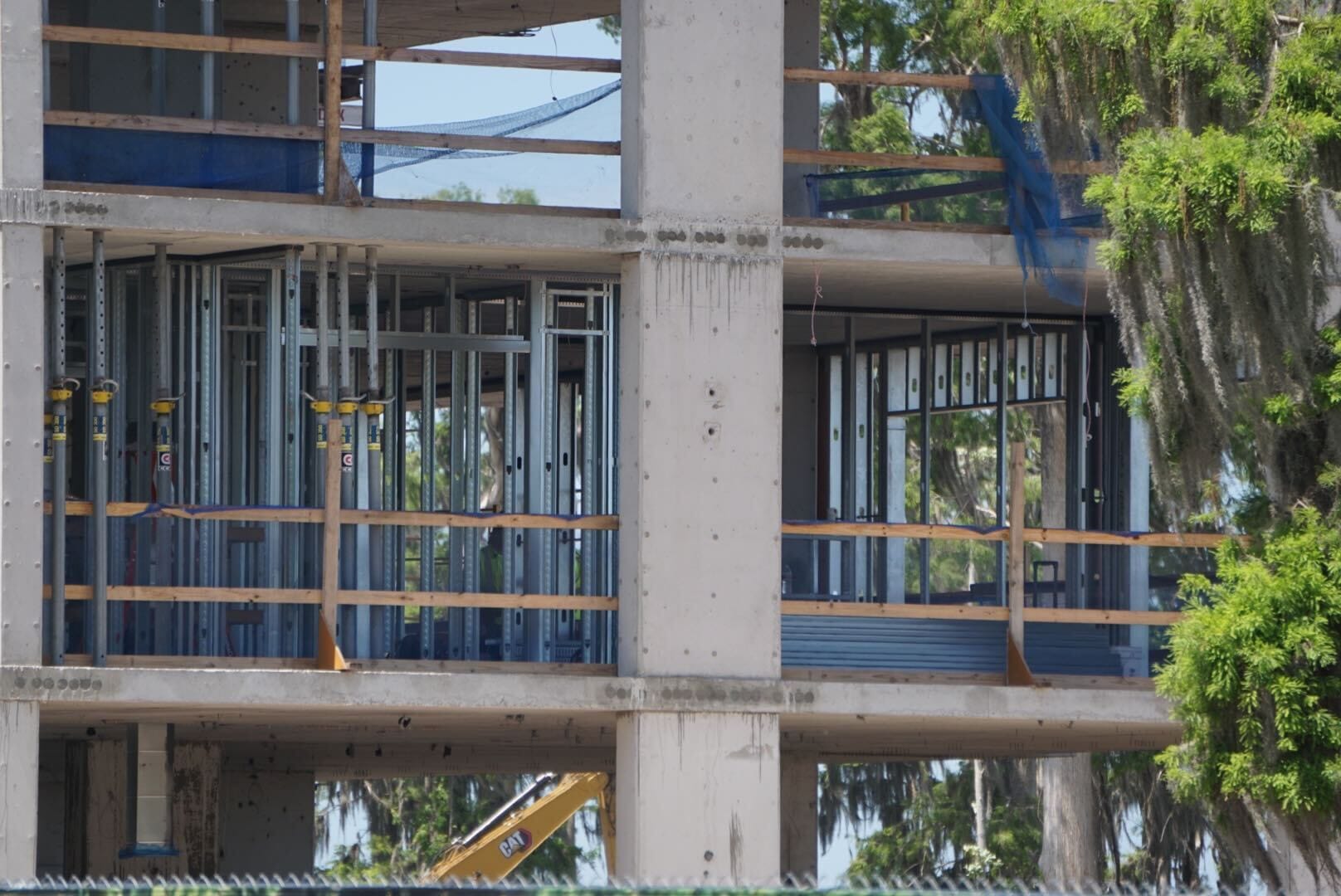 A partially constructed building with exposed concrete and steel framing is seen beside trees and construction equipment.