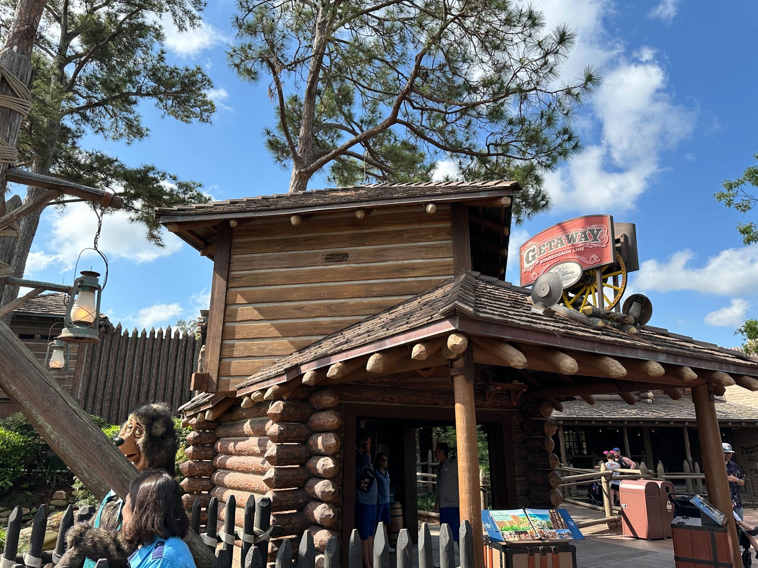Frontier-style log cabin with a "Getaway" sign, people outside, a lantern, trees, and a bright blue sky.