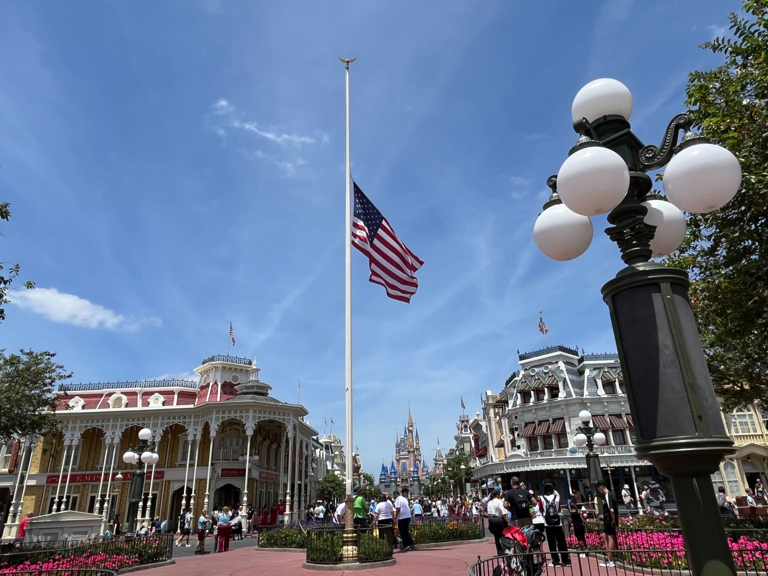 An American flag at half-mast in a busy theme park plaza, with a castle and clear blue sky in the background.