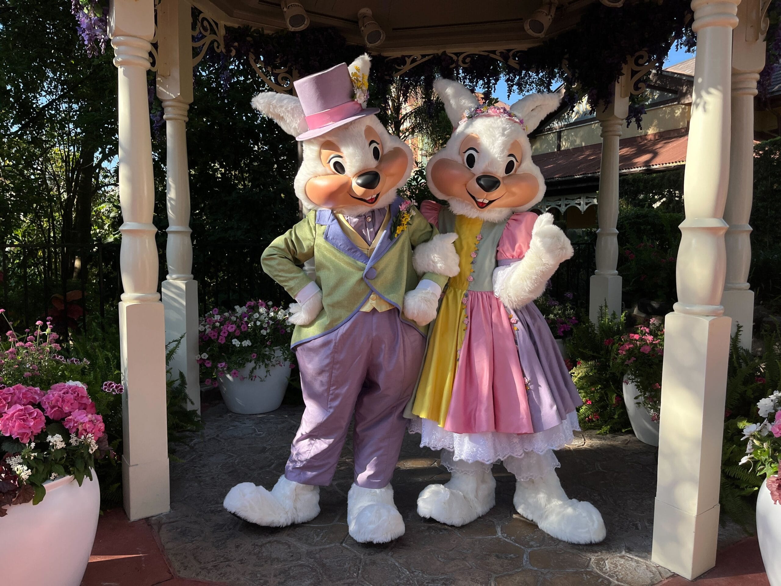 Two Easter bunnies in pastel outfits stand side by side in a garden setting with flowers and a gazebo.