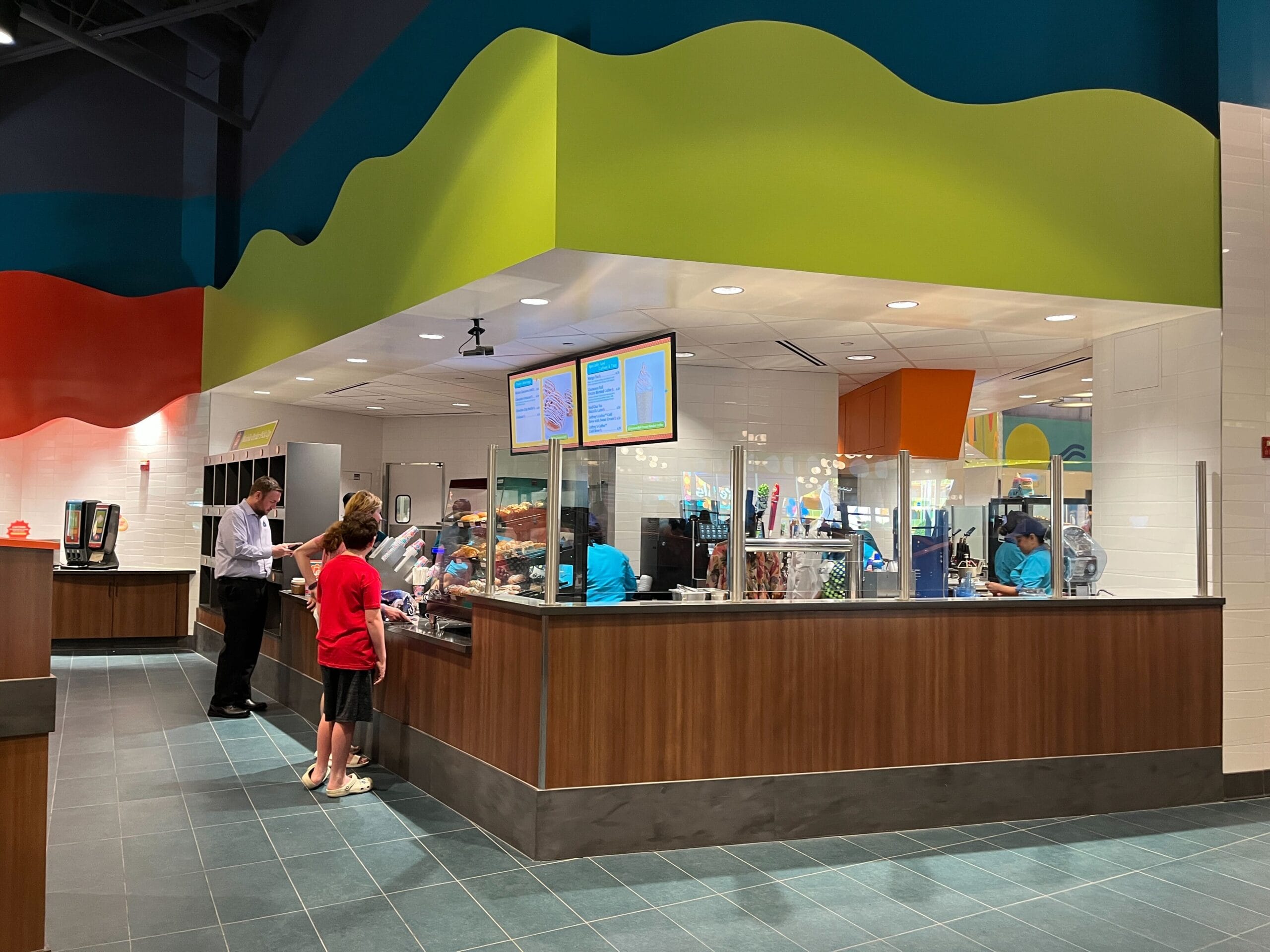 Two people stand at a counter in a modern food court. The interior features colorful wave designs and digital menu screens.