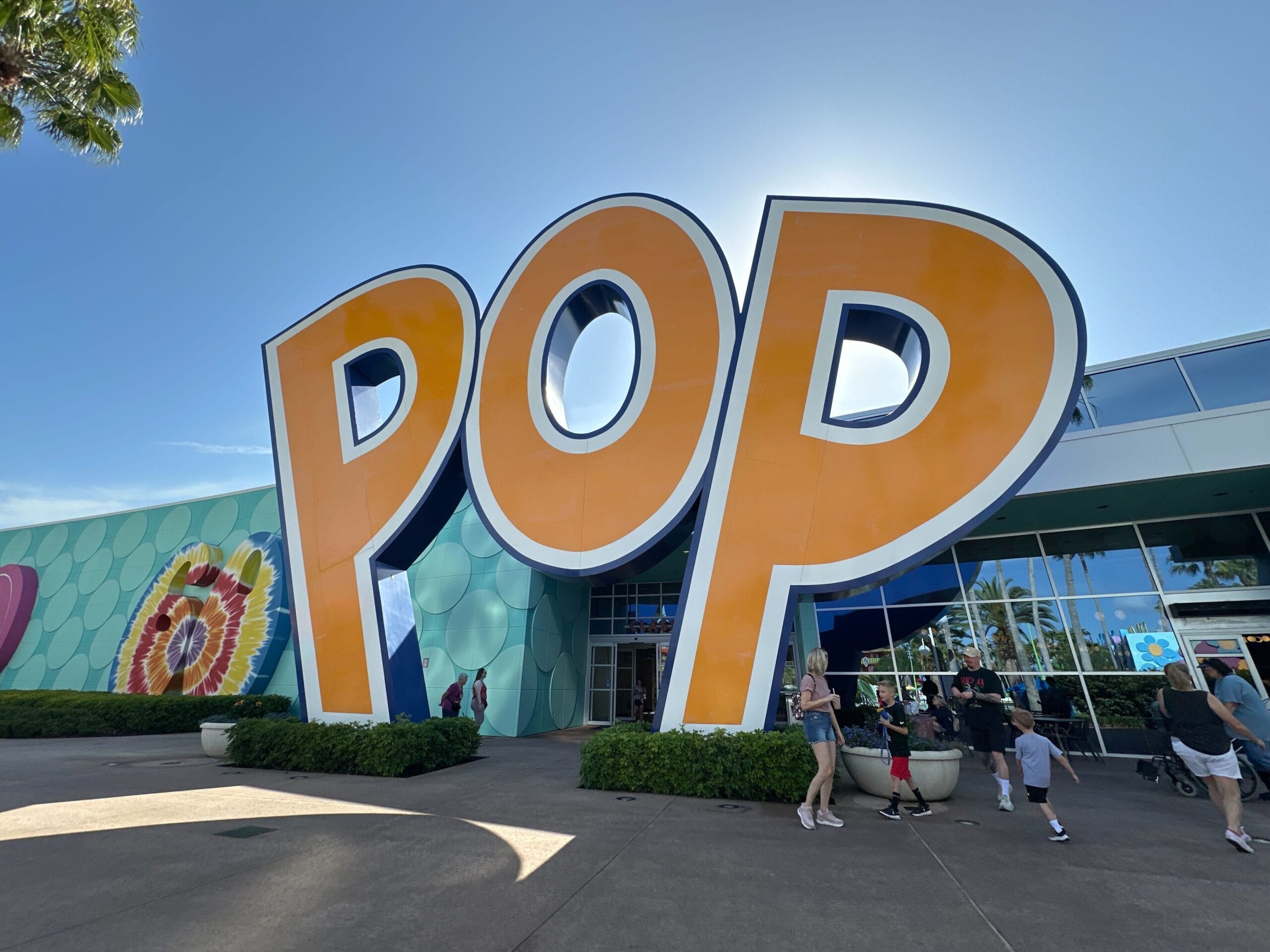 Large orange "POP" sign at an outdoor entrance with people walking by, under a clear blue sky.