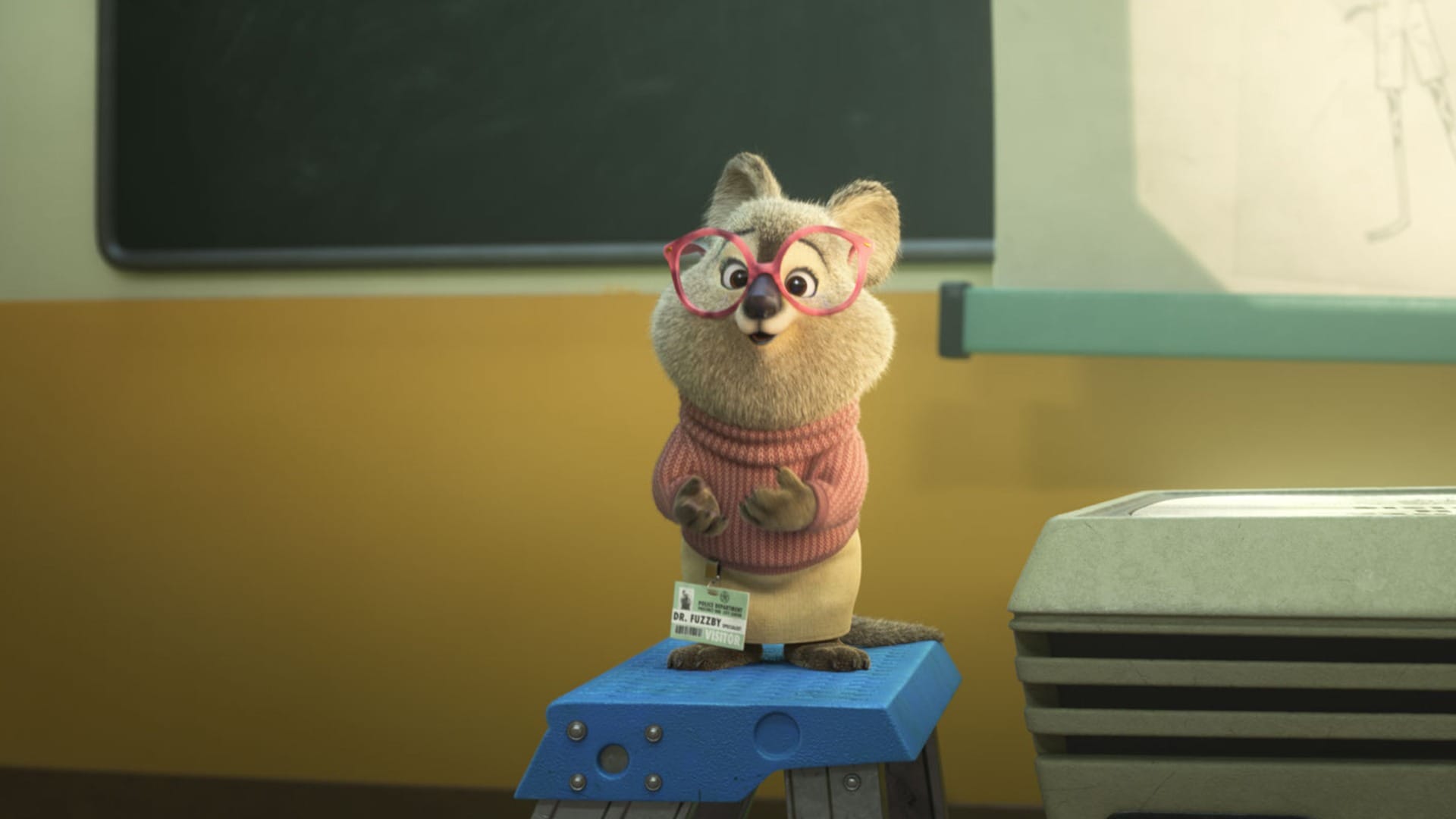 A small animated creature with large glasses stands on a stool wearing a pink sweater and name tag beside a projector. A chalkboard and yellow wall are in the background.