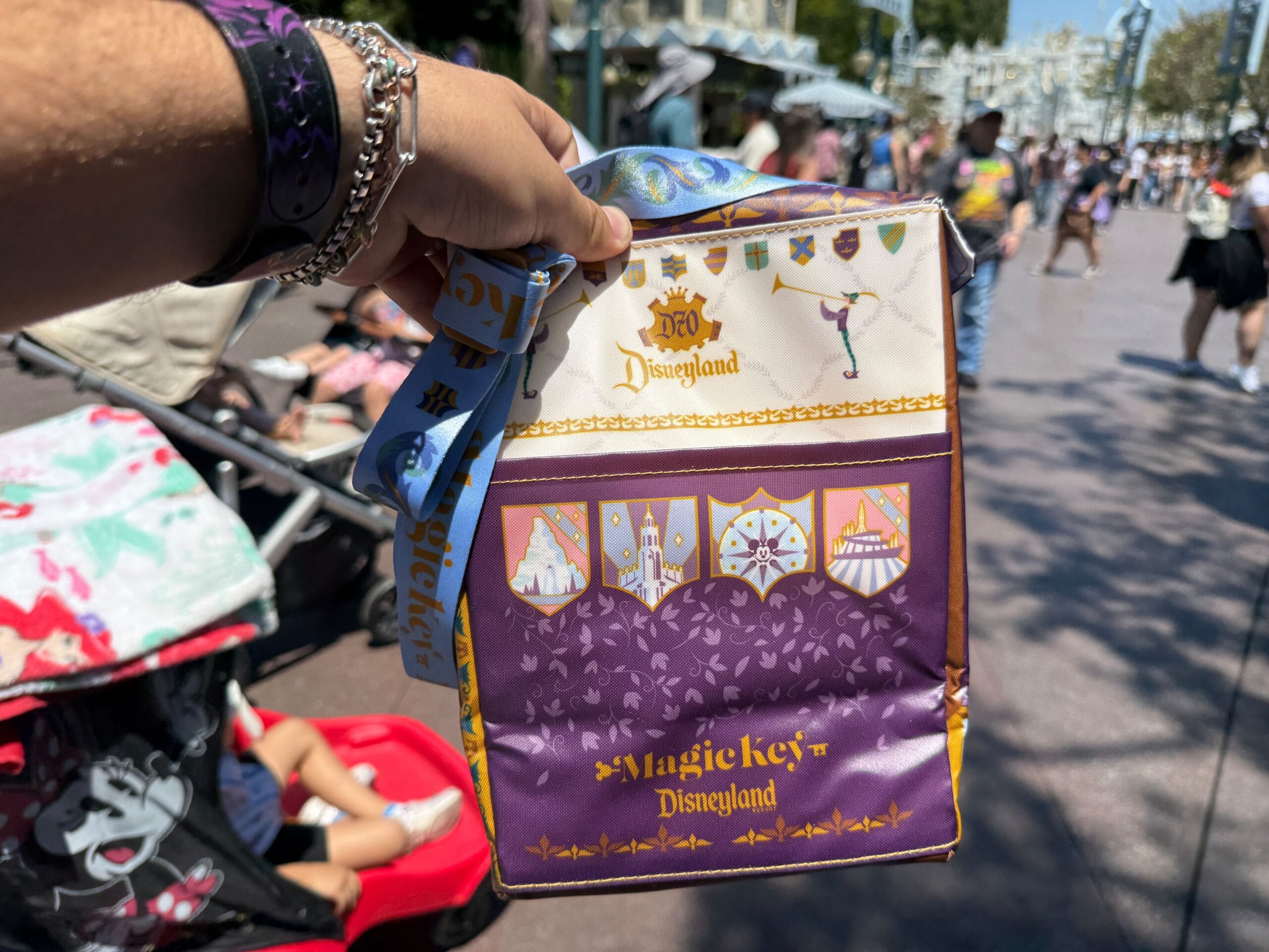 A hand holds a Magic Key Exclusive cooler bag in a crowded outdoor area at Disneyland’s 70th Anniversary.