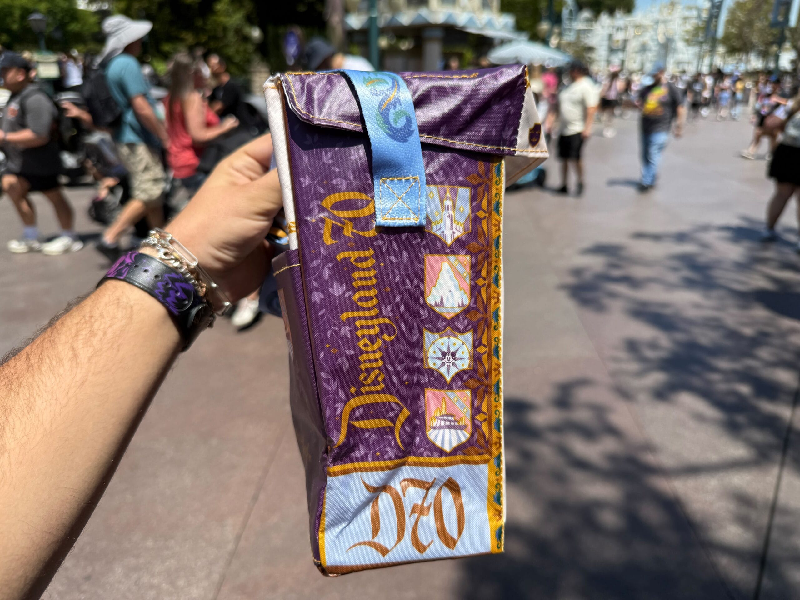 A hand holds a Magic Key Exclusive Disneyland 70th Anniversary popcorn bucket in a crowded outdoor theme park area.