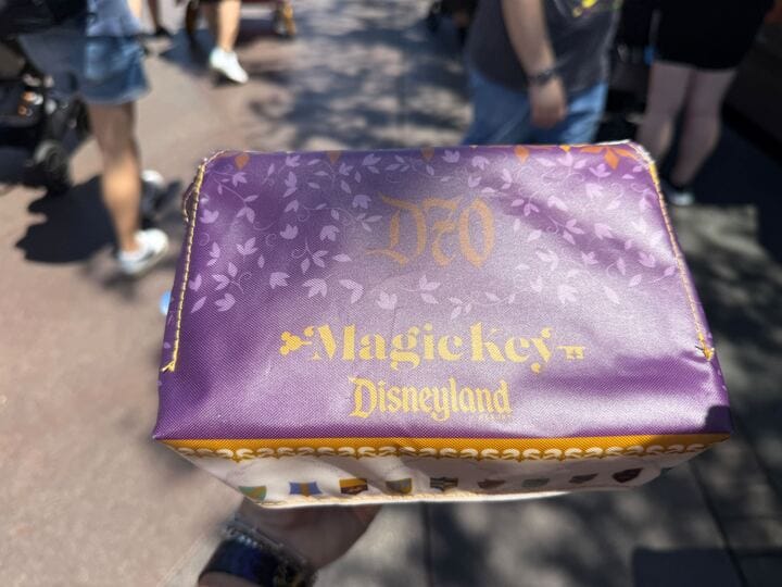 A hand holds a purple Magic Key Exclusive box with gold lettering, outdoors among people at Disneyland's 70th Anniversary.