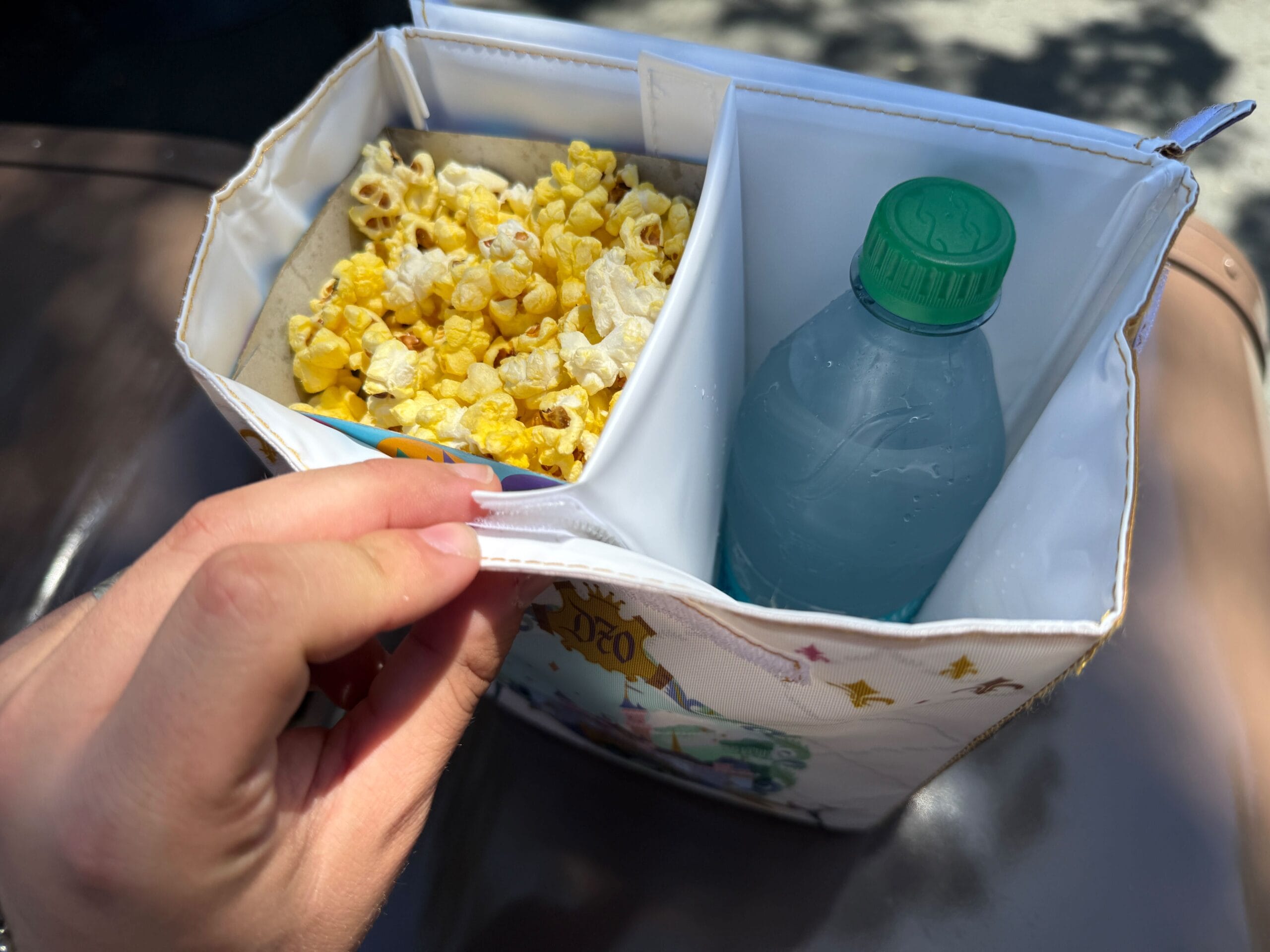 A hand holds a Disneyland 70th Anniversary cooler bag with popcorn on one side and water on the other, separated by a divider.