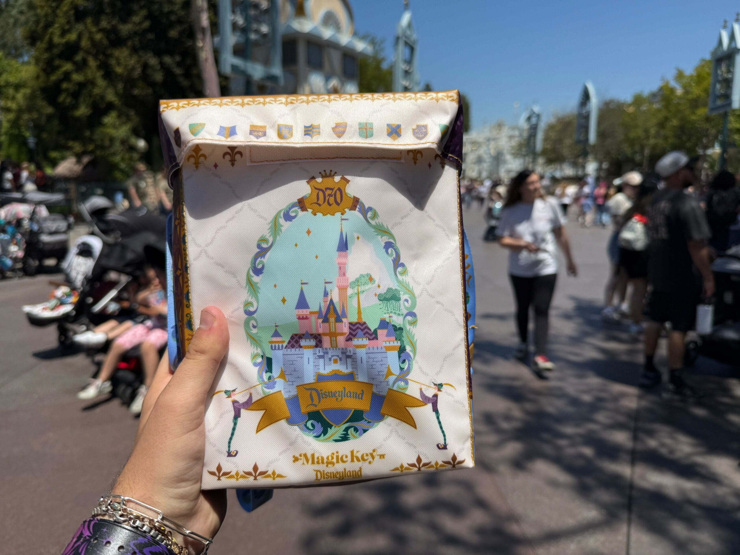 A hand holds a Magic Key Exclusive Disneyland 70th Anniversary treat bag near Sleeping Beauty Castle in a busy park walkway.