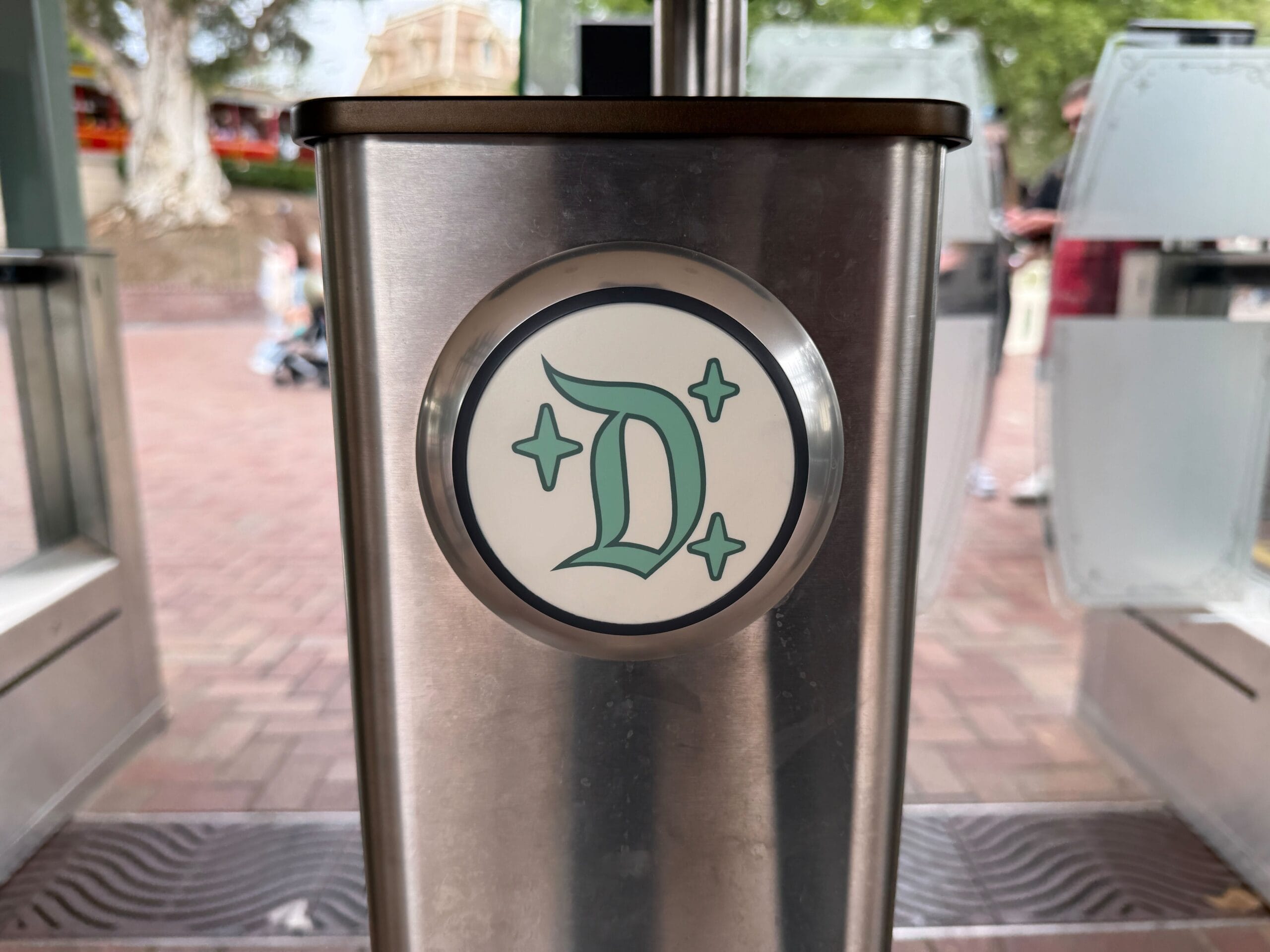 A metallic tapstile with a green "D" emblem and sparkles stands at the entrance to Disneyland.