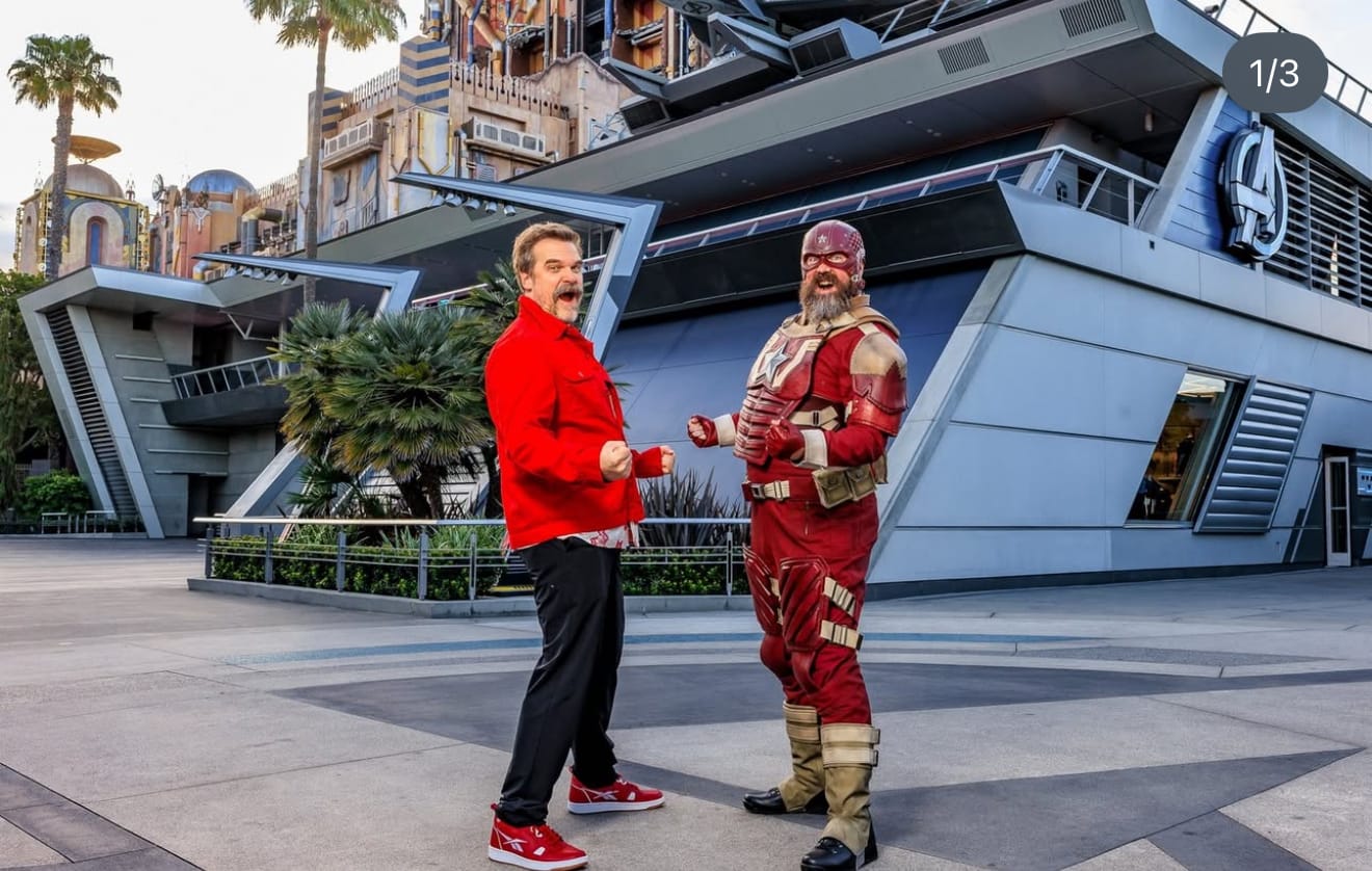 A man in a red jacket poses playfully with Red Guardian outside a modern building at Avengers Campus.