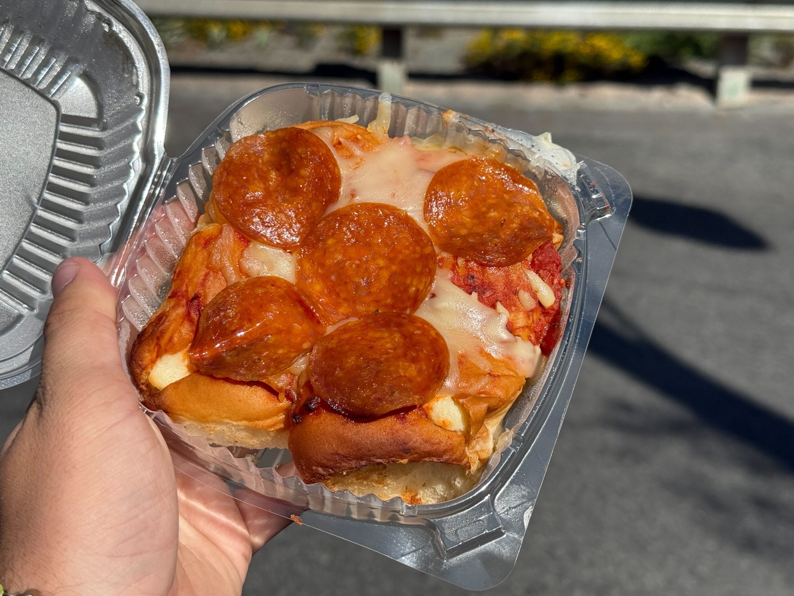 A hand holds a clear container with a square slice of pepperoni pizza topped with melted cheese.