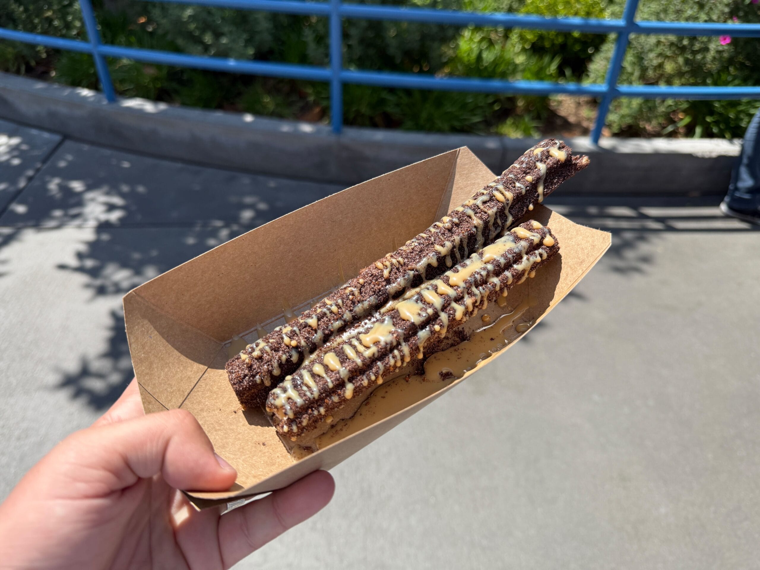 A hand holds a tray with two chocolate churros from the Churro Cart in Hollywood Land, drizzled with chocolate sauces.
