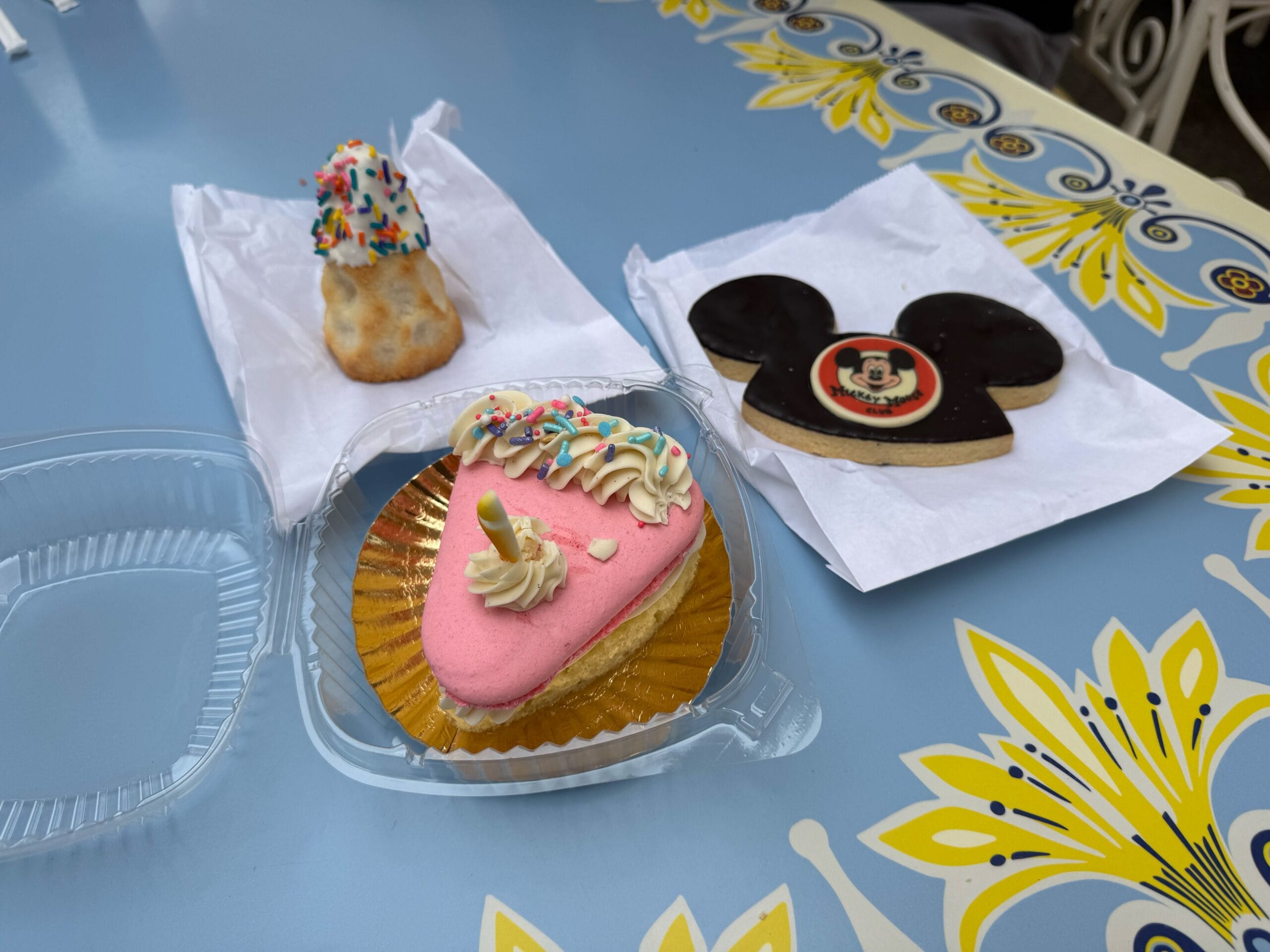 Three decorated desserts on a blue floral table: a pink cake, Matterhorn Cream Puff, and a Mickey Ear Hat Cookie.