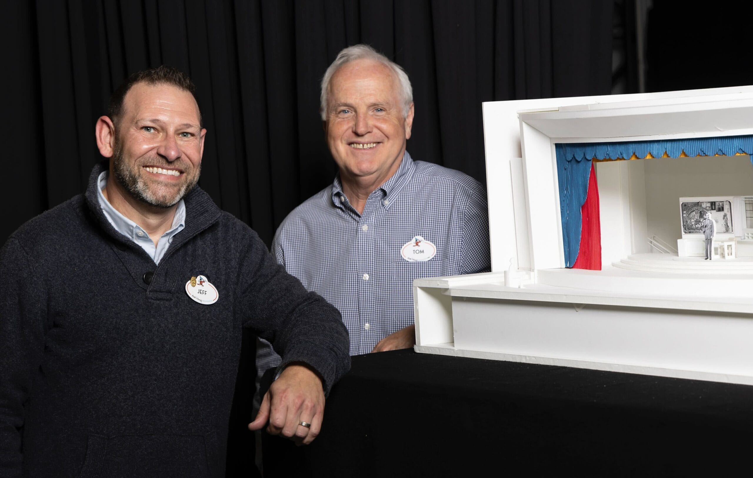 Two men smiling beside a small white model of a theater stage, inspired by Disney attractions and Audio-Animatronic magic.